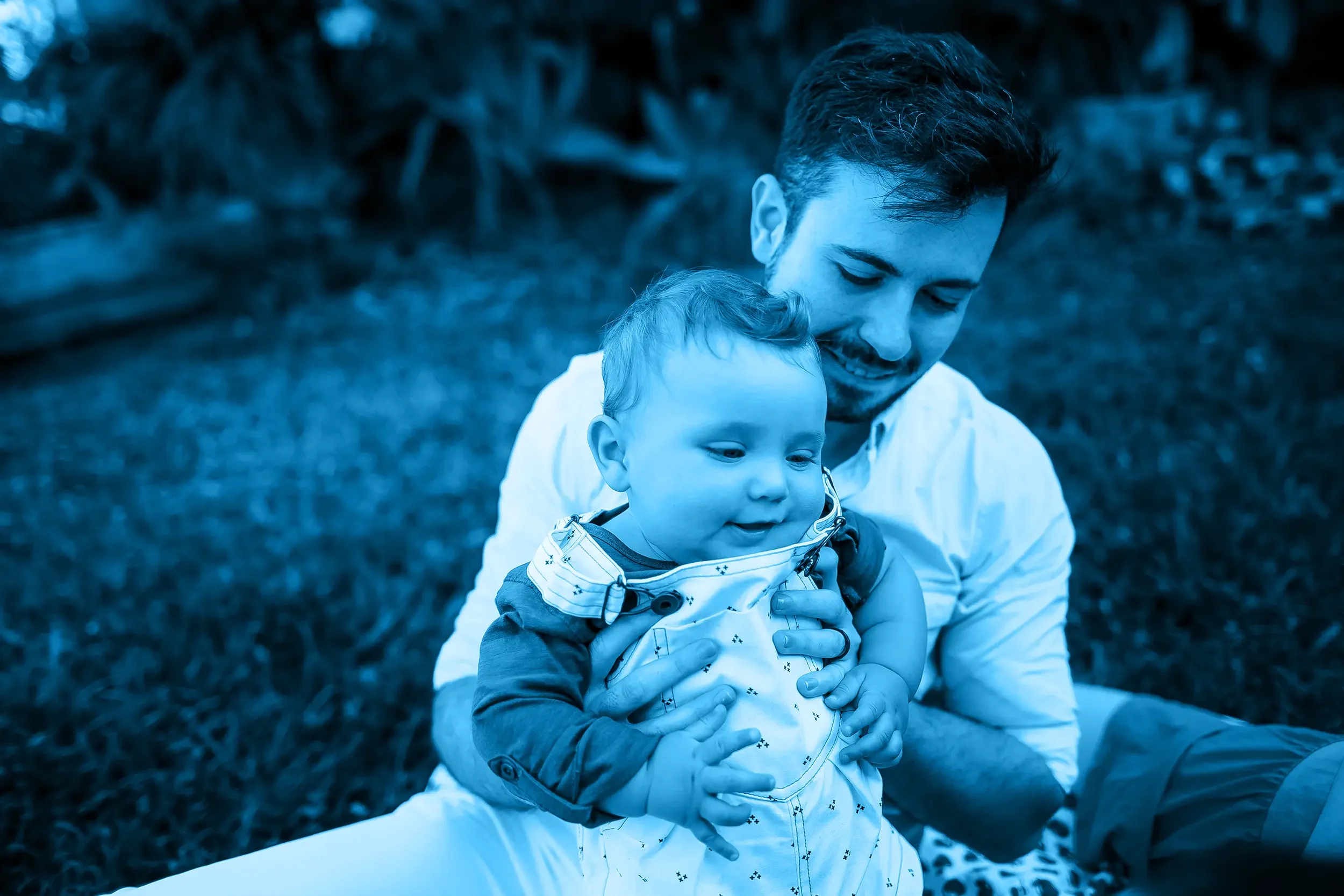 A father holding a young child outdoors, both smiling, with trees and grass in the background.