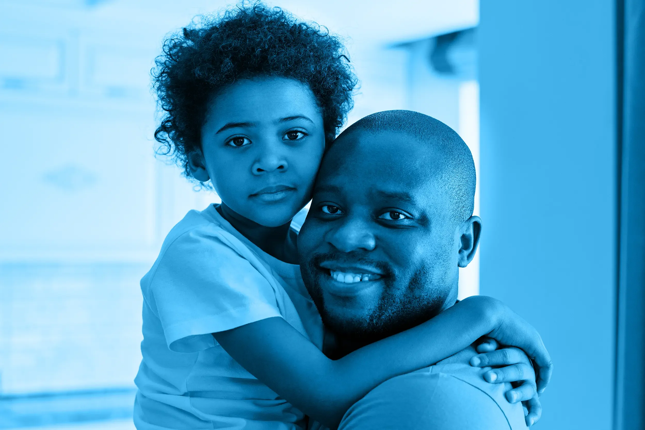 A young girl with curly hair embraces her dad, both smiling and looking at the camera, in a brightly lit indoor setting.