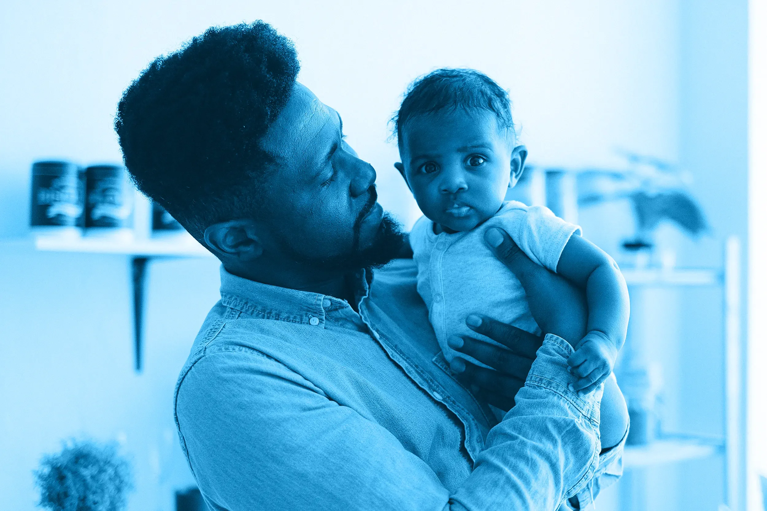 A dad holding a baby in a room with shelves and cans in the background, both looking at the camera.