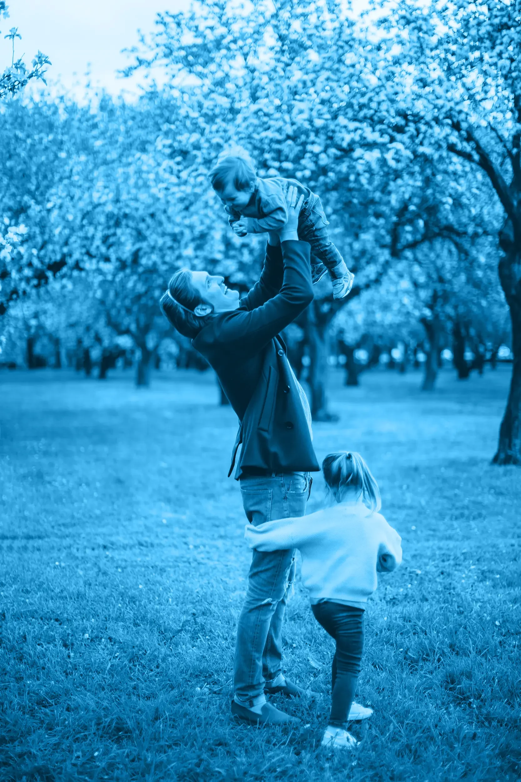 A dad playing with two children in a park with flowering trees, with a blue color overlay.