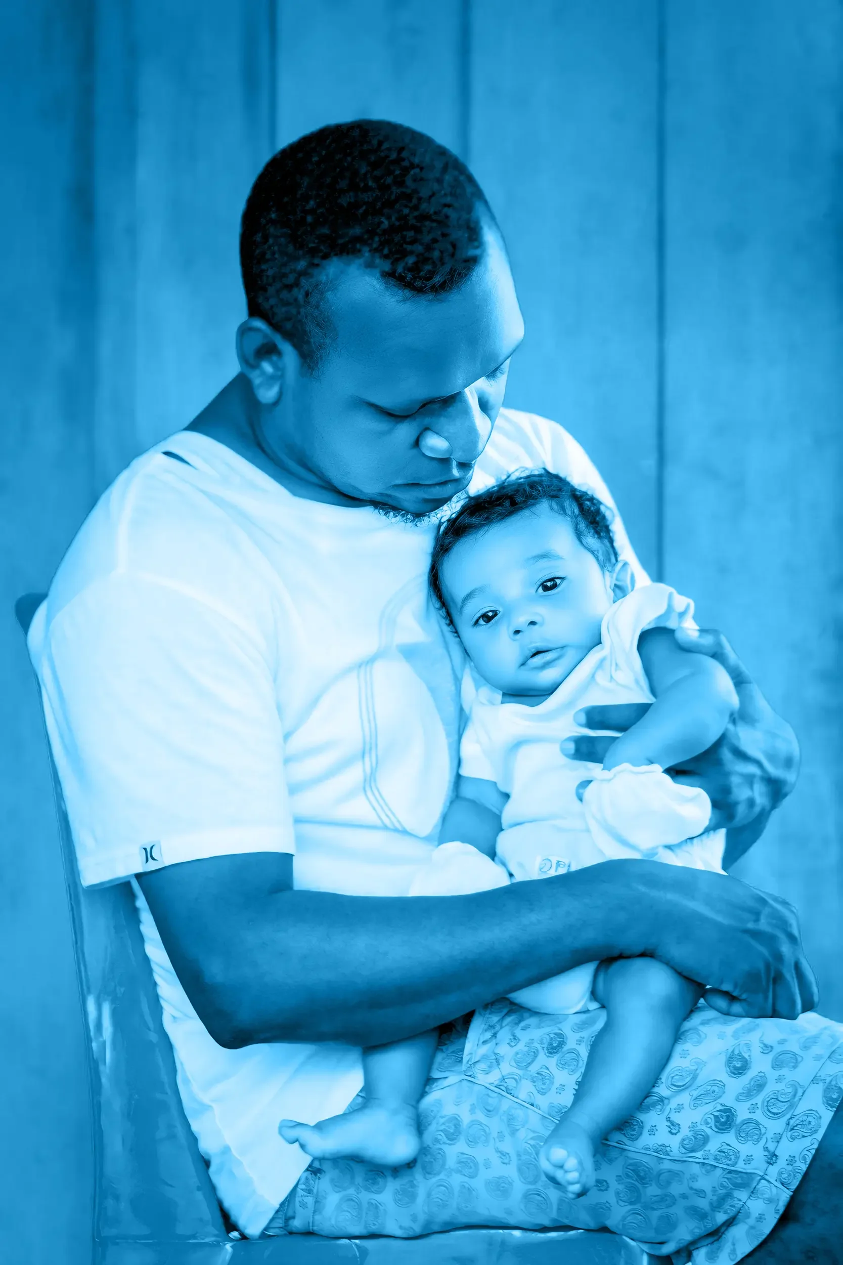 A dad with short curly hair, wearing a white t-shirt, is holding a young child with curly hair and light skin. The man is looking down at the child, who is sitting in his arm and making eye contact with the camera.