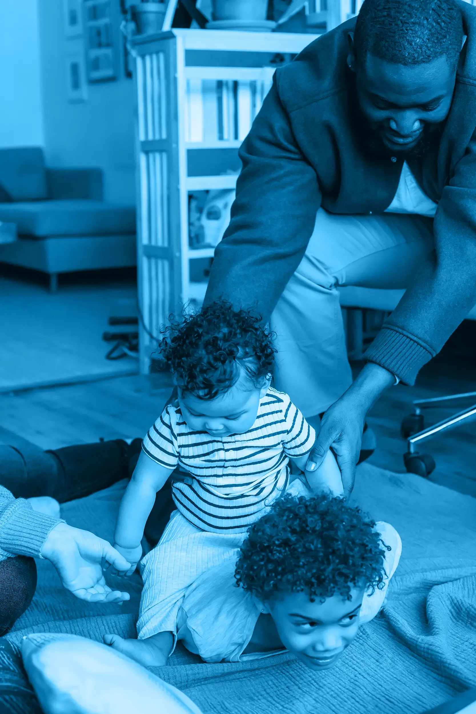 A dad and two children on a blanket indoors, engaged in a playful activity. The man is leaning over, holding one child's hand, while the children are on their stomachs, playing and looking down.