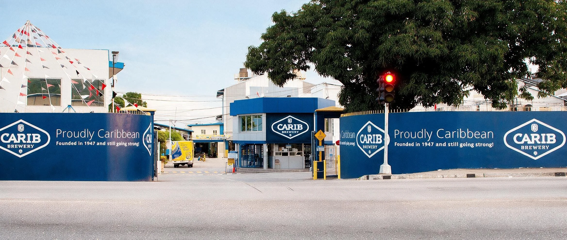Entrance of Carib Brewery with blue advertising banners, a security booth, and a red traffic light, with a large tree and buildings in the background.
