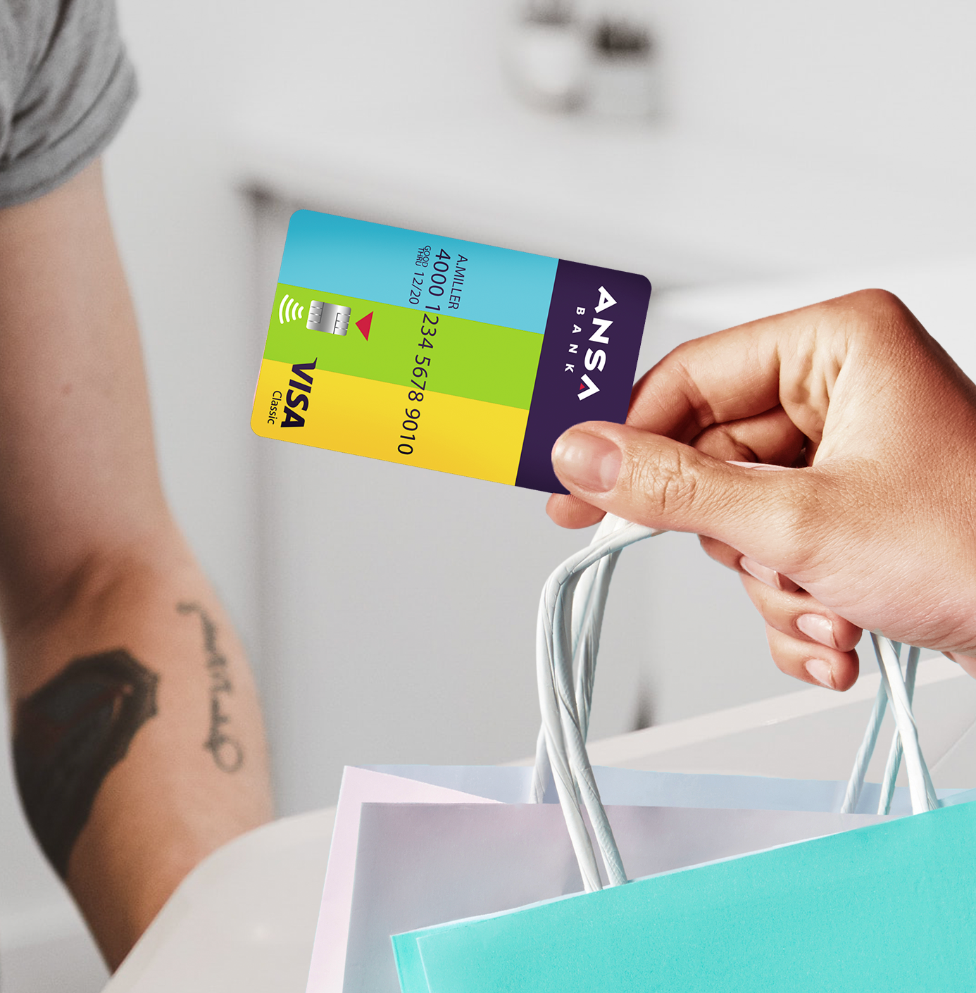 A hand holding a colorful ANSA BANK Visa debit card with electronic payment symbols and a shopping bag with paper and plastic handles.