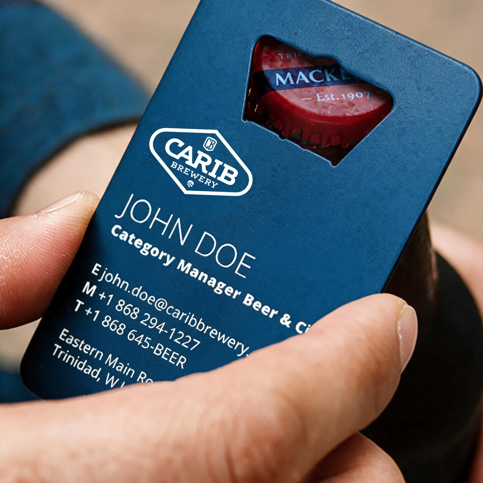 Business card holder with a bottle cap visible through a cutout on the card, featuring the Mackeson logo. The card displays contact details for John Doe, a category manager at Carib Brewery in Trinidad.