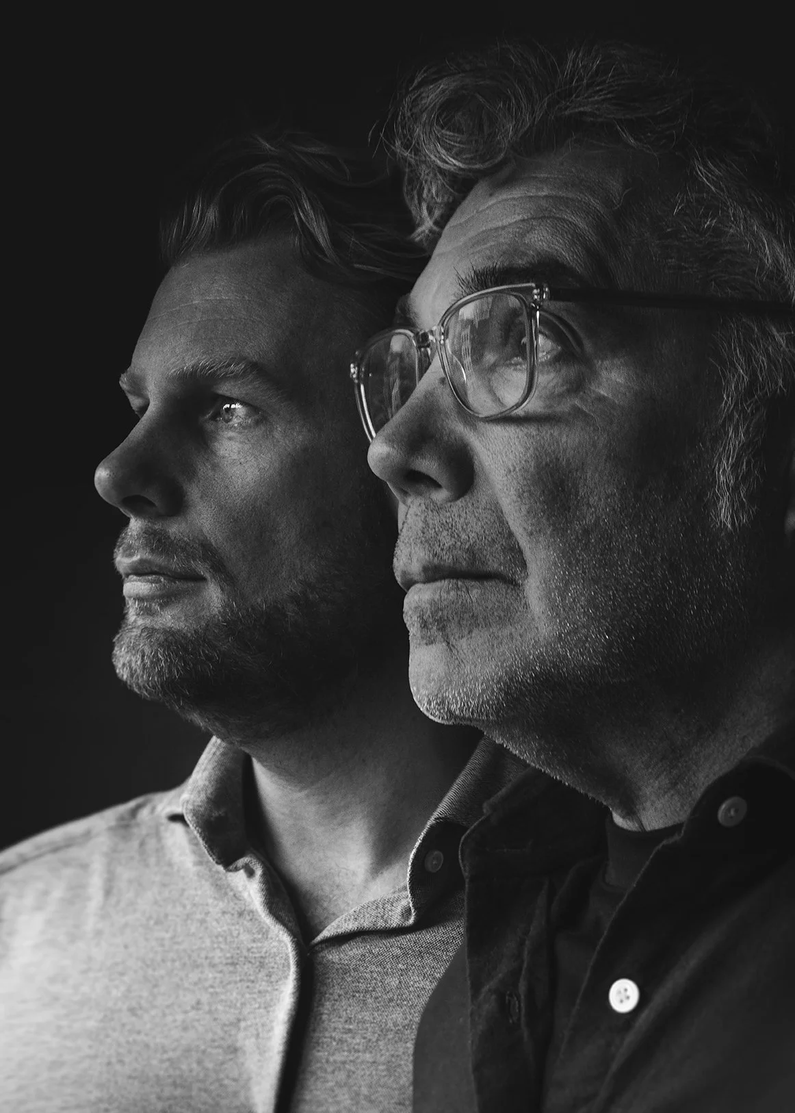 Black and white portrait of two men facing left, one wearing glasses and the other with facial hair, both looking into the distance.
