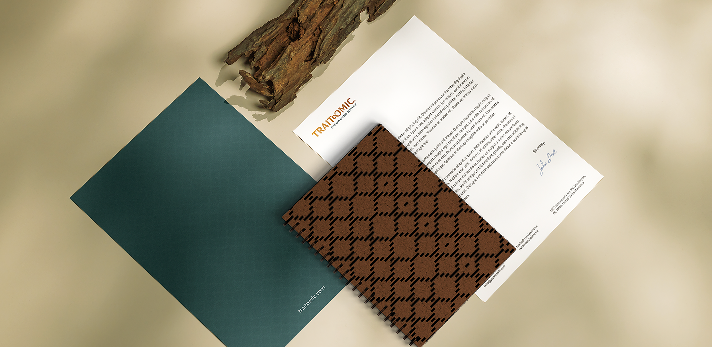 An arrangement of Traitomic stationery including a piece of driftwood, a sheet of teal paper, a paper with 'Traitomic' written on it, a brown patterned fabric, and a printed letter with a signature, all on a plain light-colored surface.