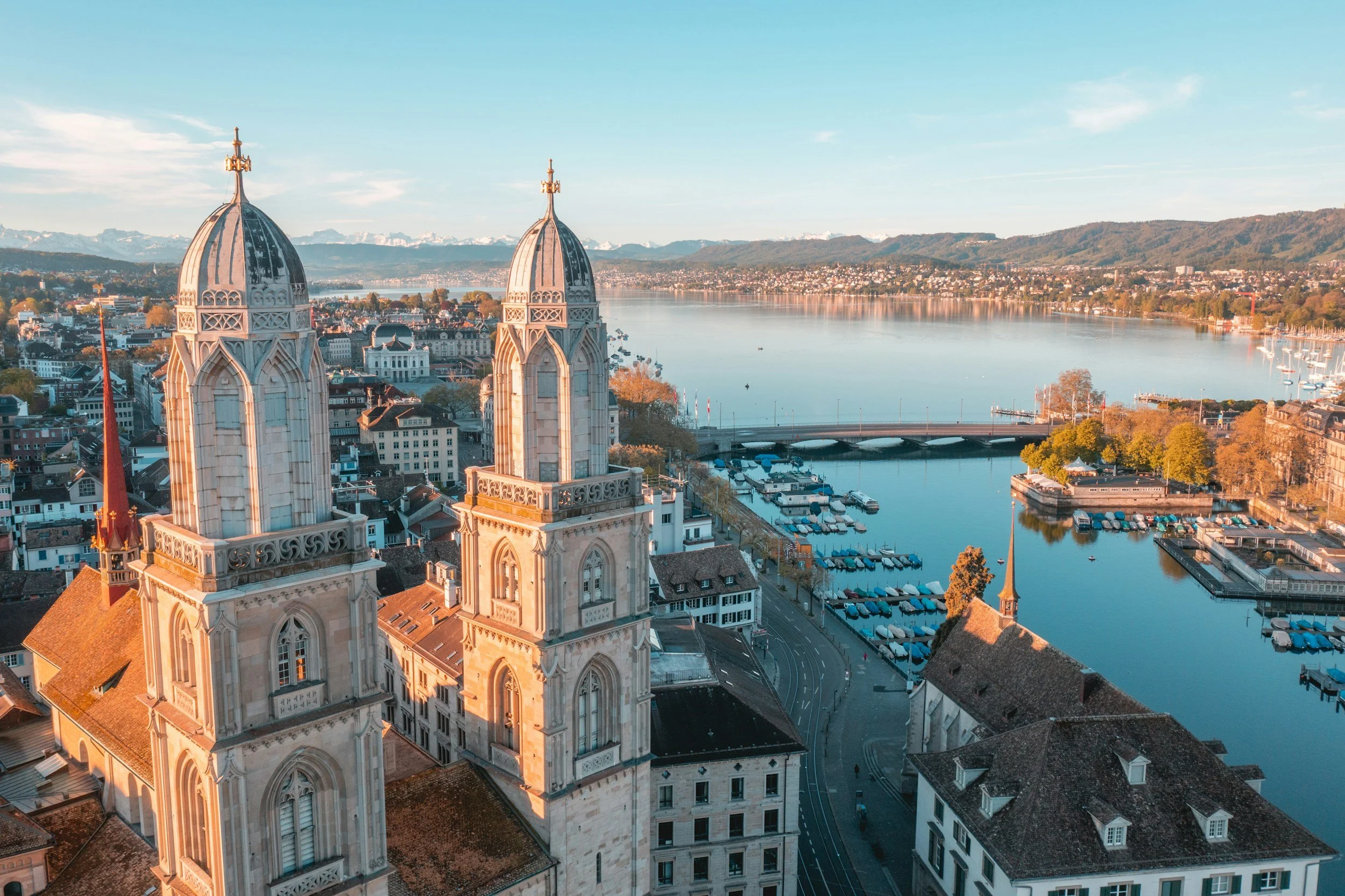 Aerial view of Zurich's historic church towers, a river, boats, and mountains in the distance during sunset.