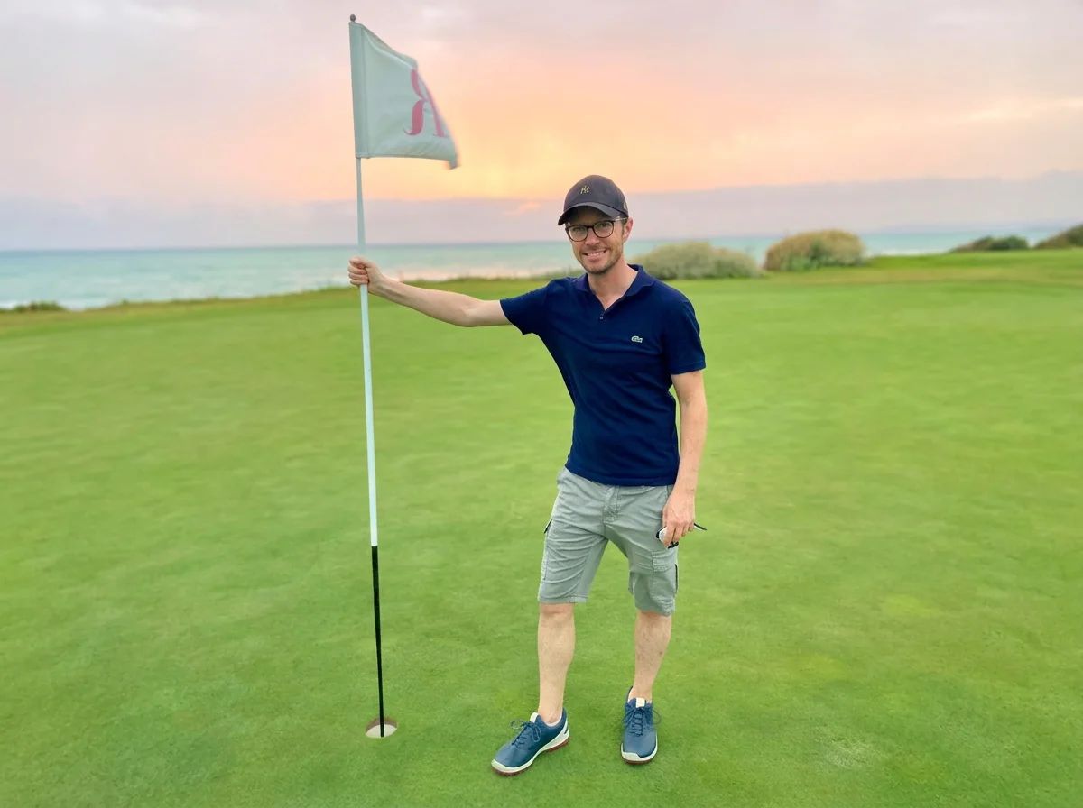 Man standing on a golf green holding a golf flagstick near the hole, with a sunset over the ocean in the background.