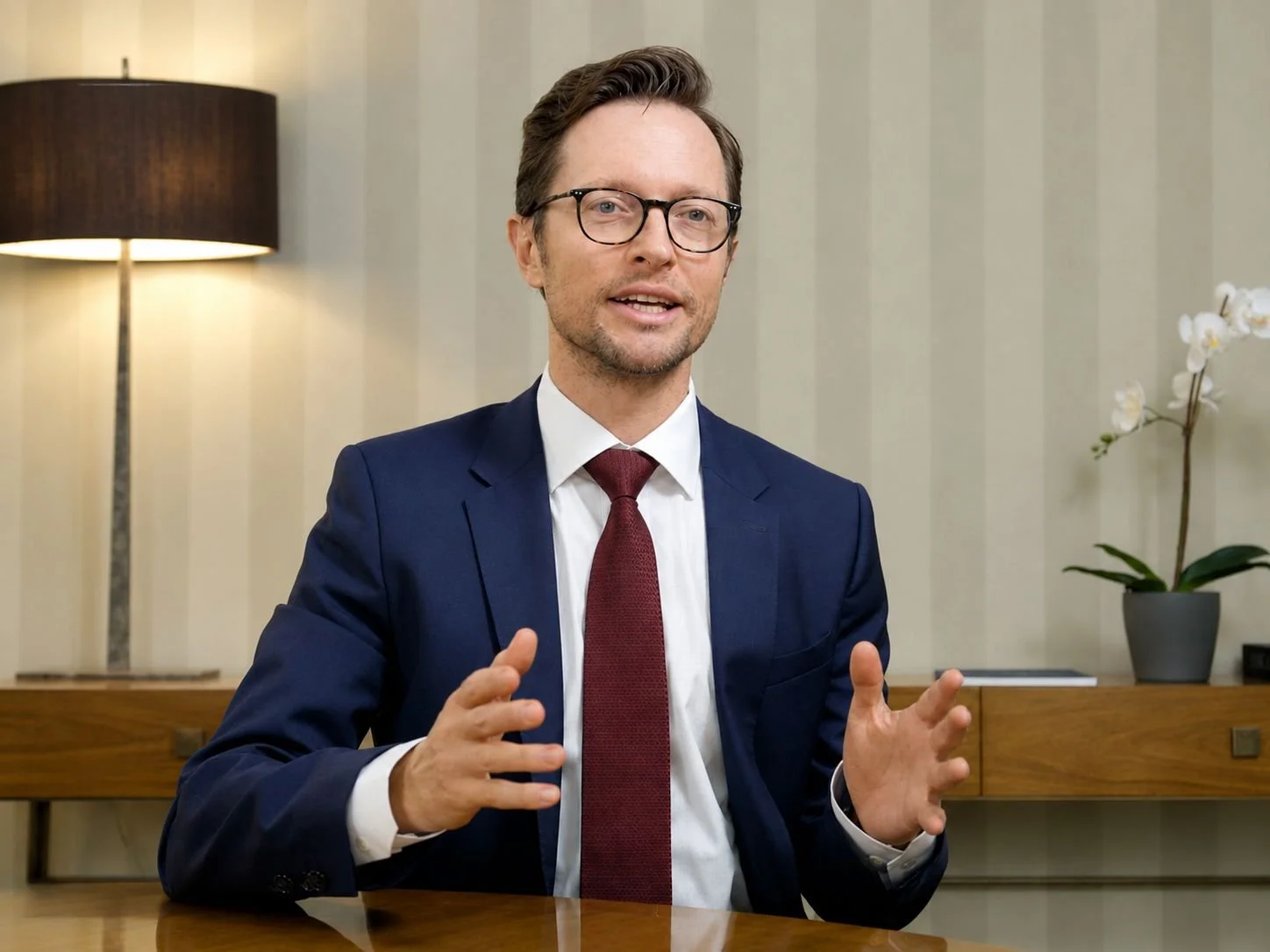 A man wearing glasses, a navy suit, white shirt, and a red tie, speaking and gesturing with his hands in an office setting.