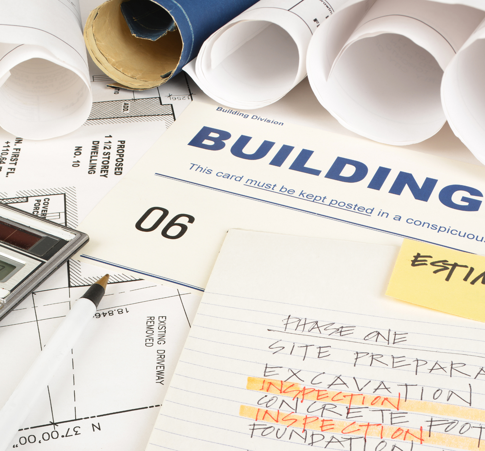 A construction blueprint, a building permit card, rolled architectural plans, a pen, and a hand-written note about site preparation and excavation work on a table.