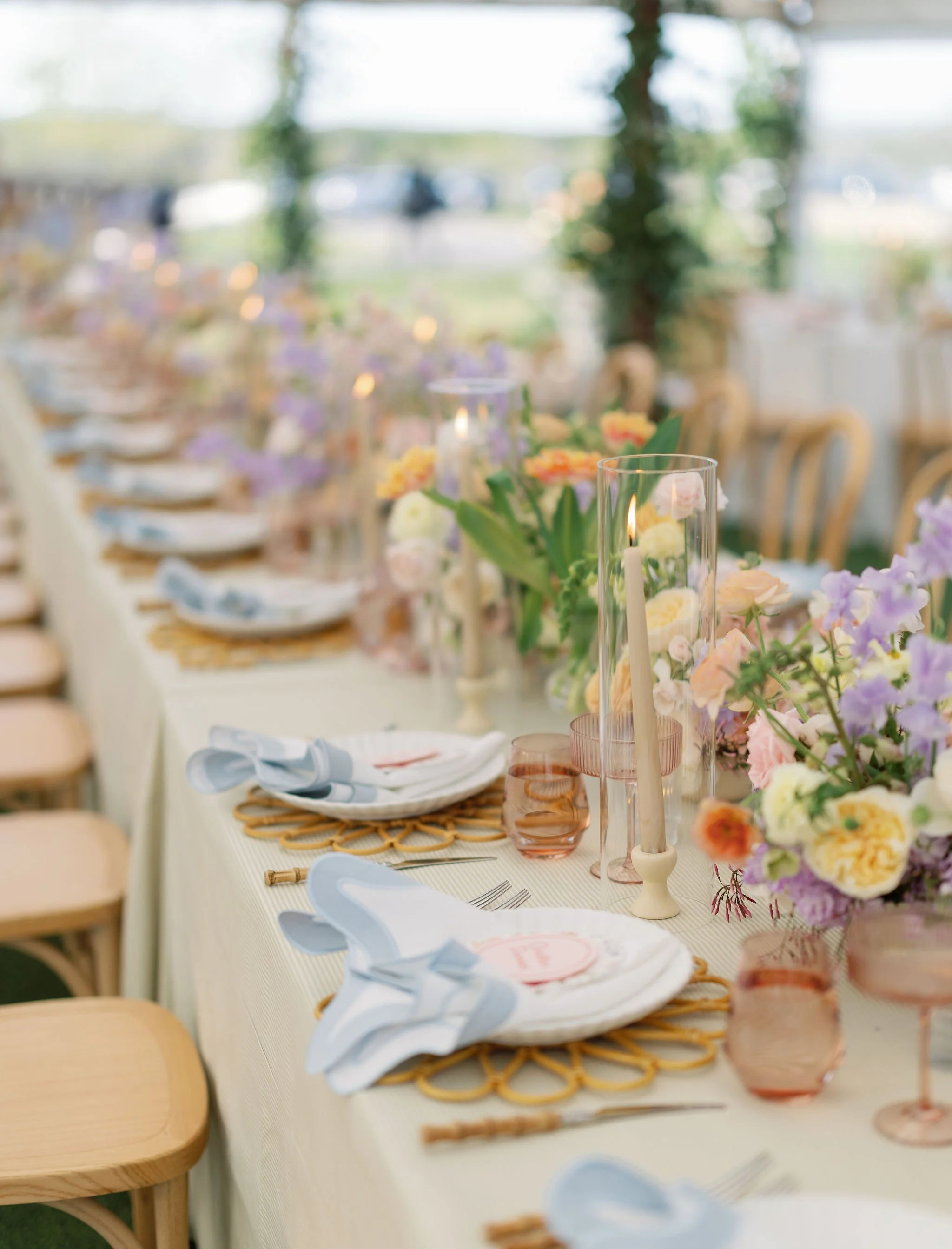 A long table decorated with pink, white, and purple flowers, candle holders with candles, and place settings with white napkins and plates, set up for a formal event or celebration.