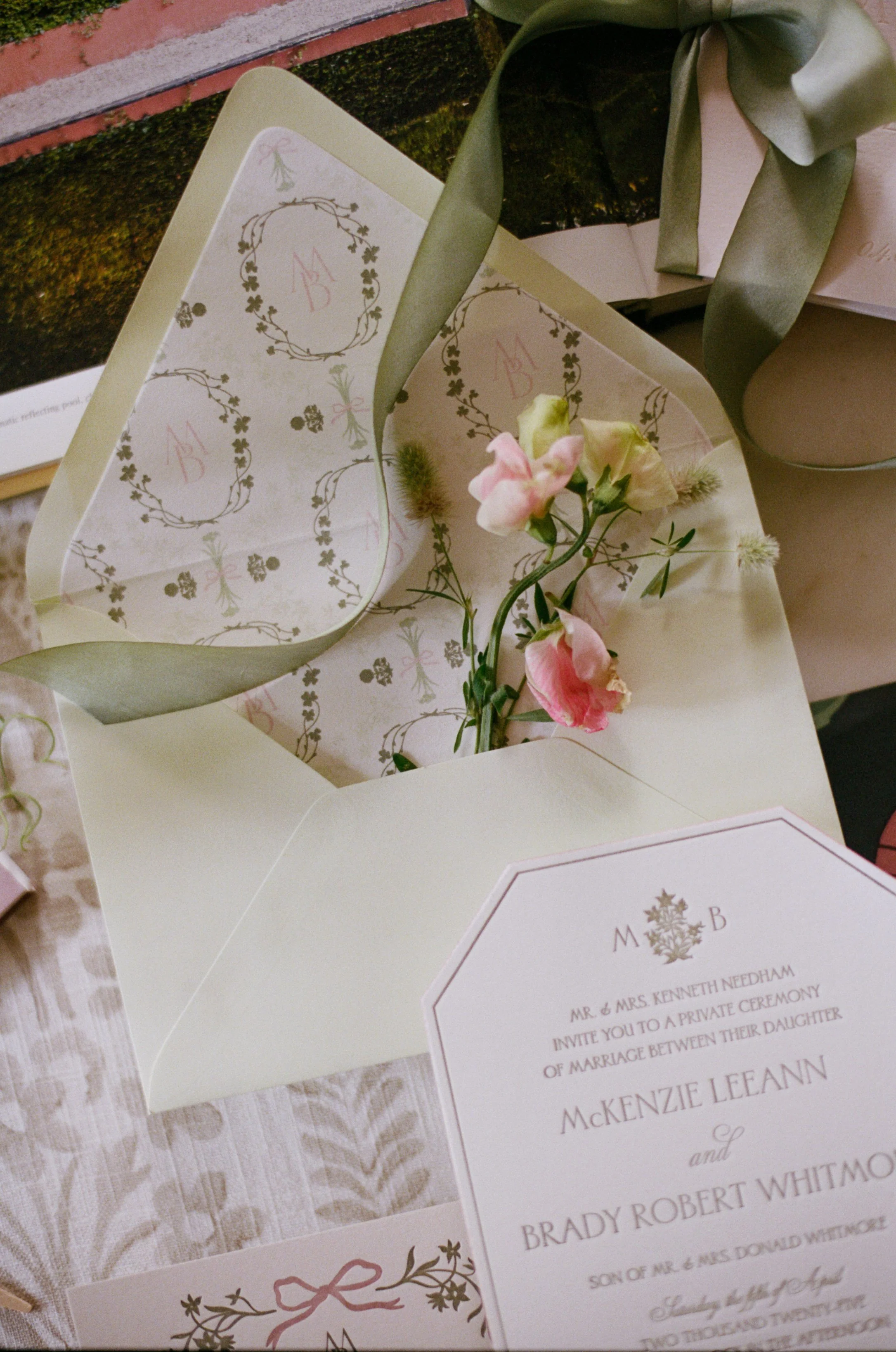 A wedding invitation featuring floral and monogram design with pink and green accents, along with a small sprig of pink flowers, all displayed on a textured surface.