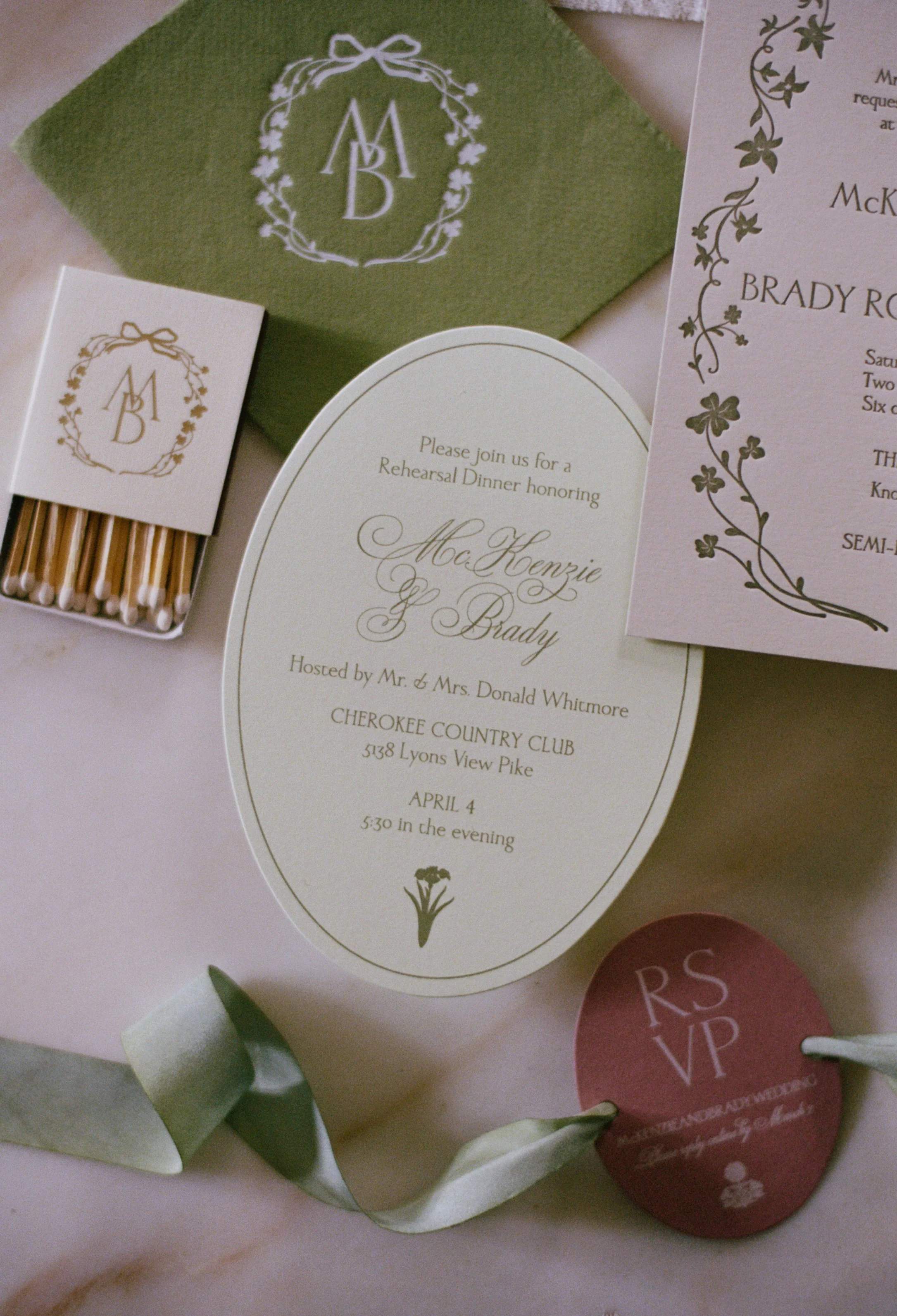 Invitation for a rehearsal dinner, a green envelope and a matchbox with a monogram, with accompanying decorative items such as a candle and flowers.