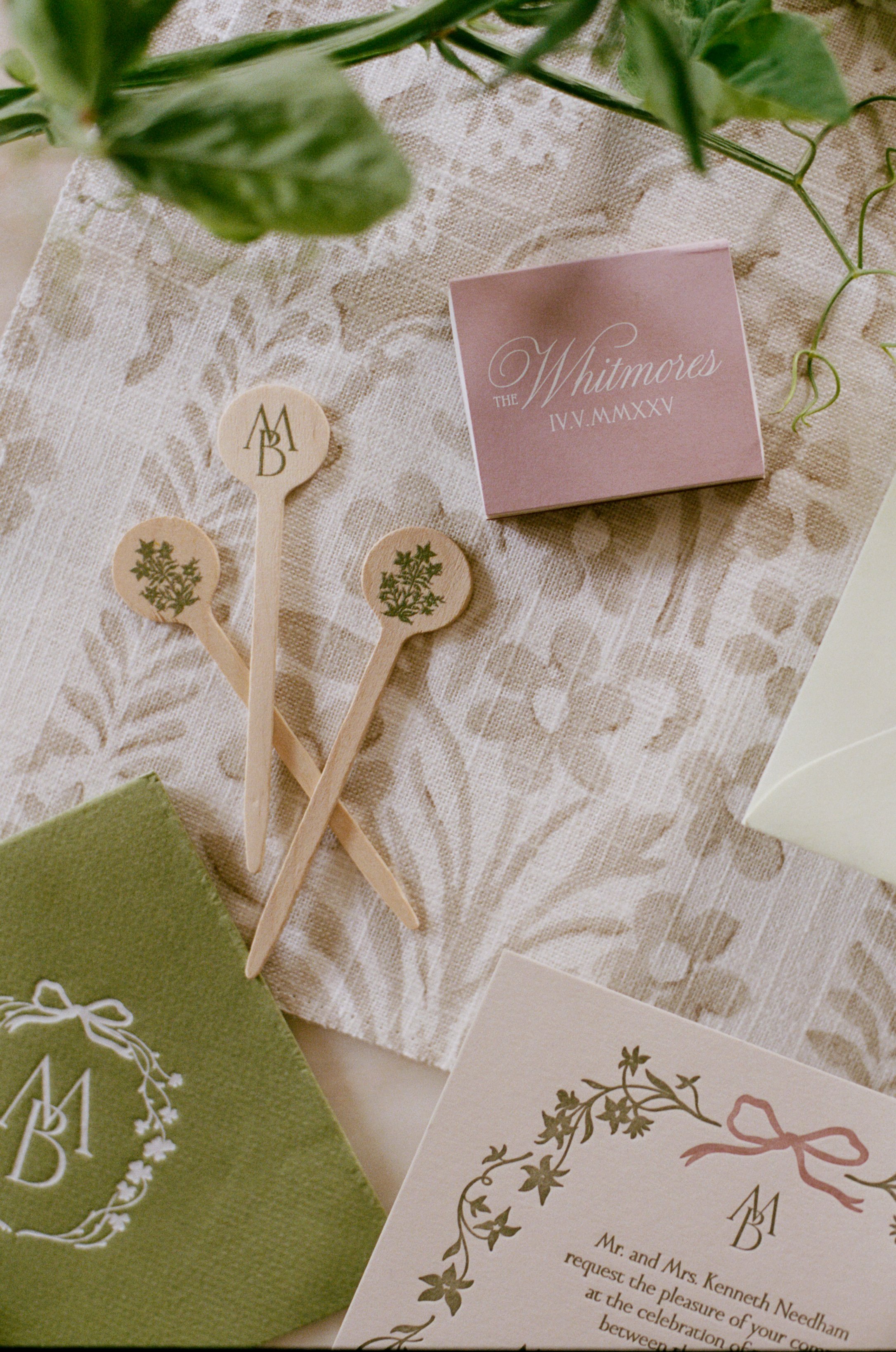Wedding invitation suite with a pink card displaying 'The Whitmores IVV. MMXXV,' olive branch illustrations, three wooden hand fans with greenery, and various other stationery items on a lace tablecloth.