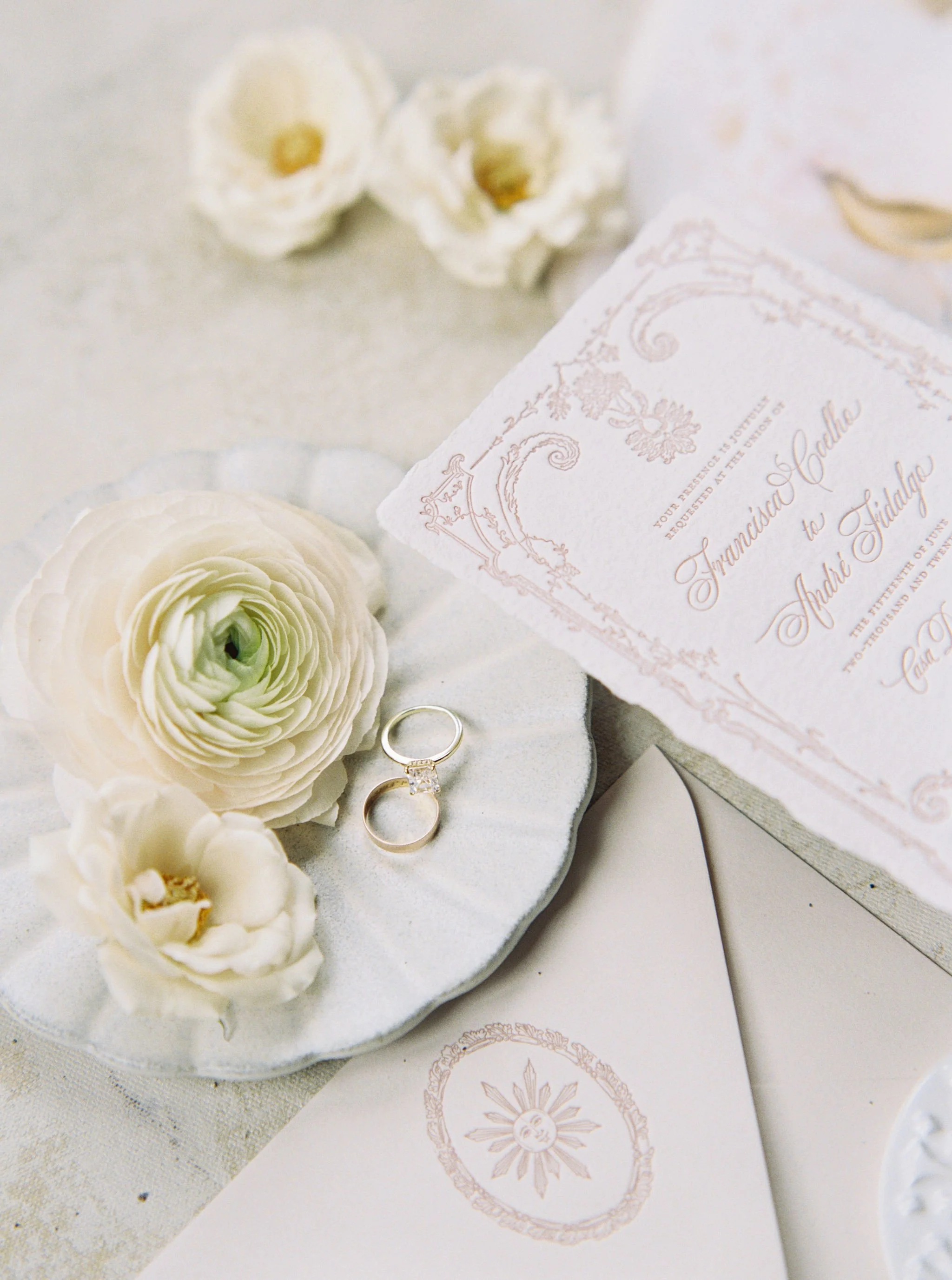 A wedding invitation card on a white plate, surrounded by white flowers and two wedding rings.