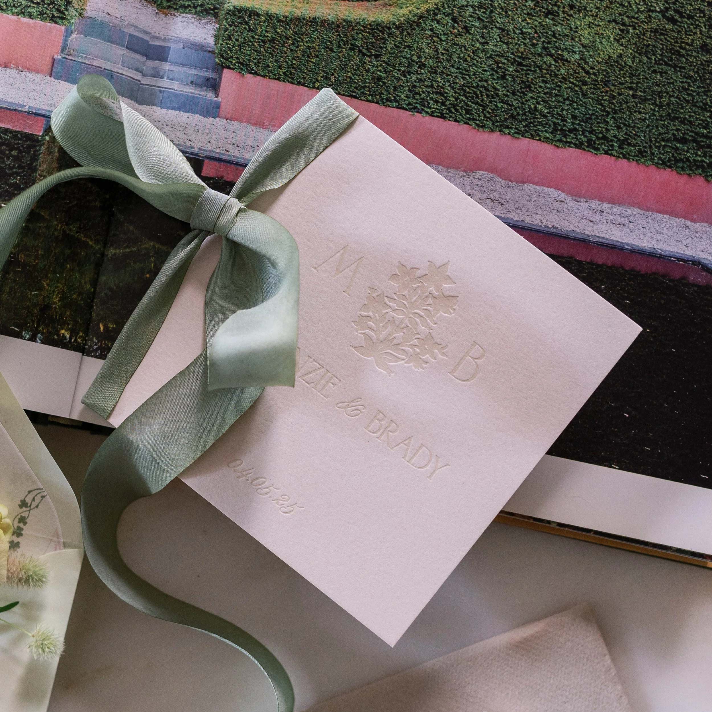 A close-up of a beige greeting card with embossed floral design and the word 'HAPPY' visible, tied with a light green satin ribbon, in front of a background featuring a photograph of stairs, shrubs, and a red wall.