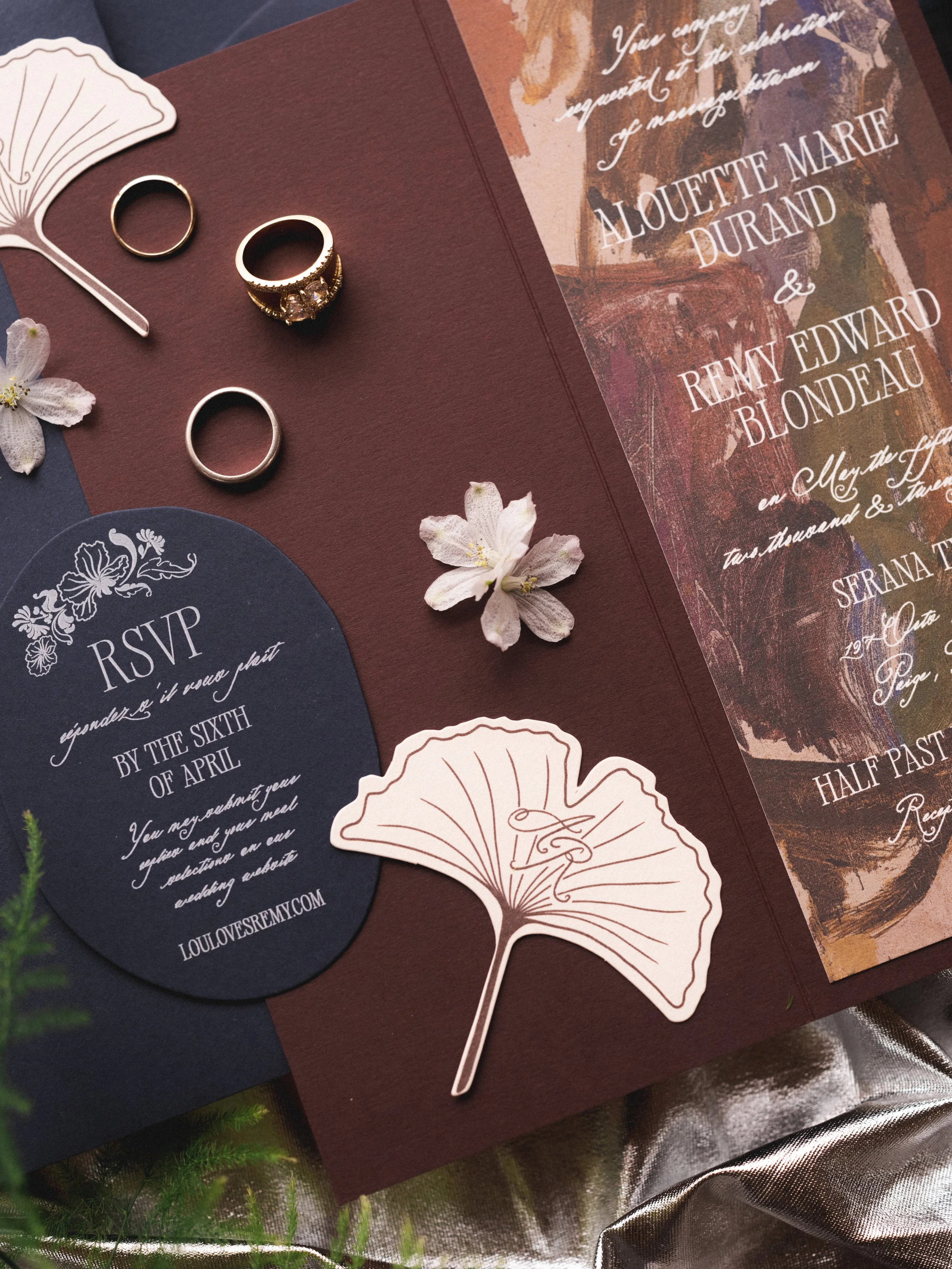 Close-up of wedding invitations, rings, and decorative paper cutouts on a textured brown and navy background with flowers and greenery.