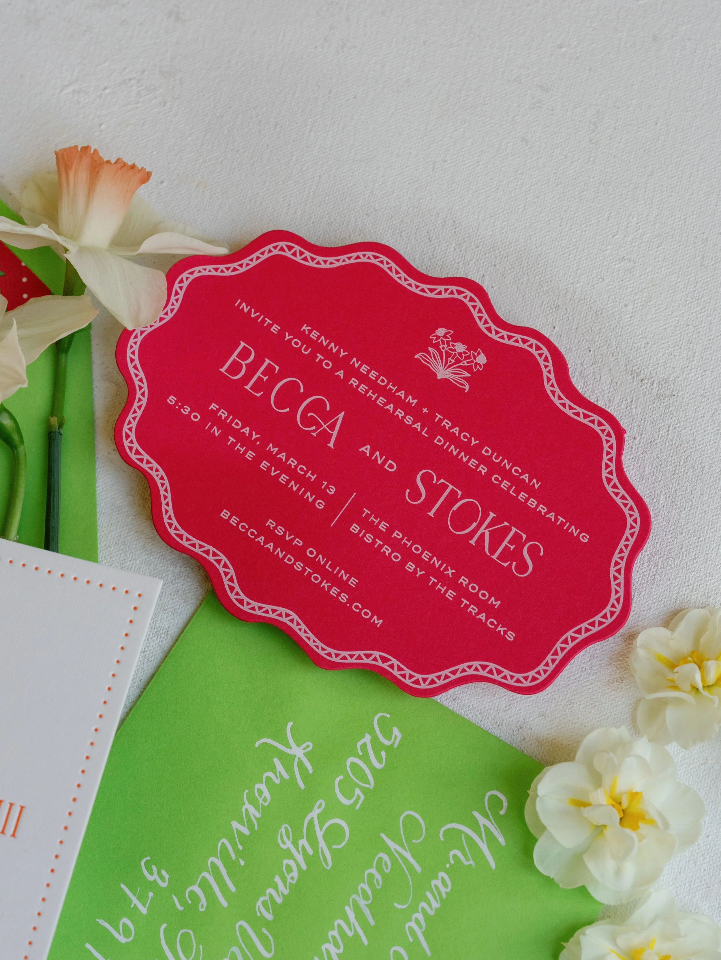 Close-up of a red scalloped edge invitation card with white text, surrounded by white flowers and green and white cards on a white surface.
