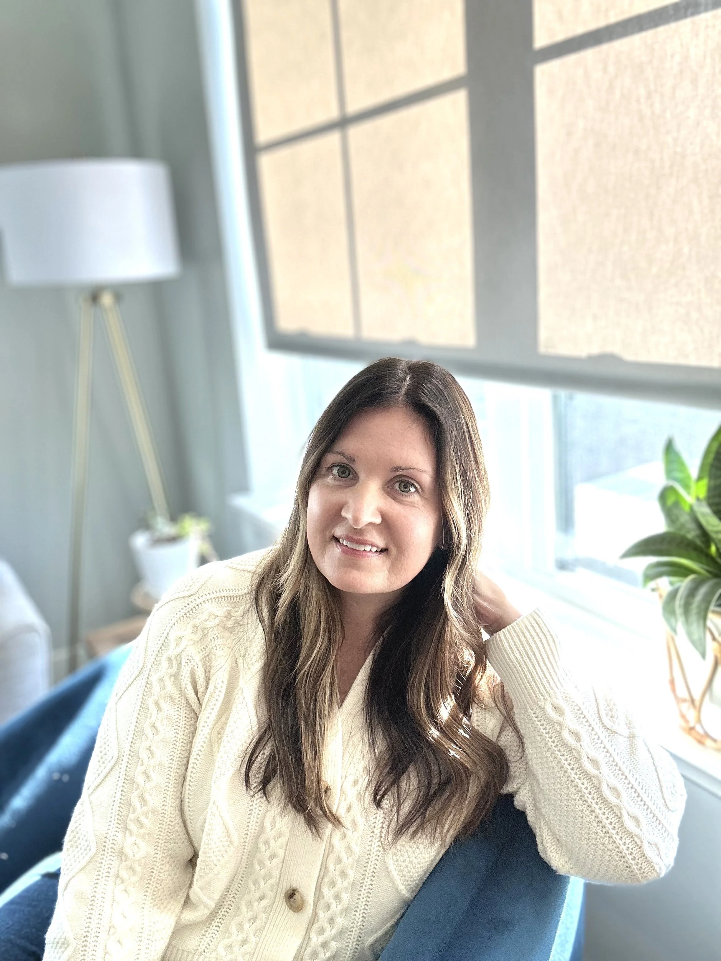 A smiling woman with long brown hair sitting on a blue chair in a room with large windows and natural light, with houseplants in the background.