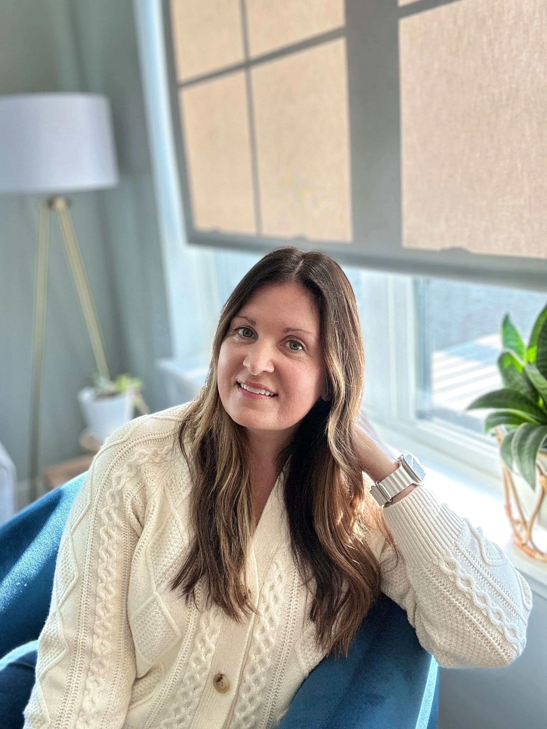 A woman mental health counselor with long brown hair, wearing a cream-colored knit sweater and a white wristwatch, sitting on a blue chair in front of a window with beige blinds, and a houseplant to her right, smiling at the camera.