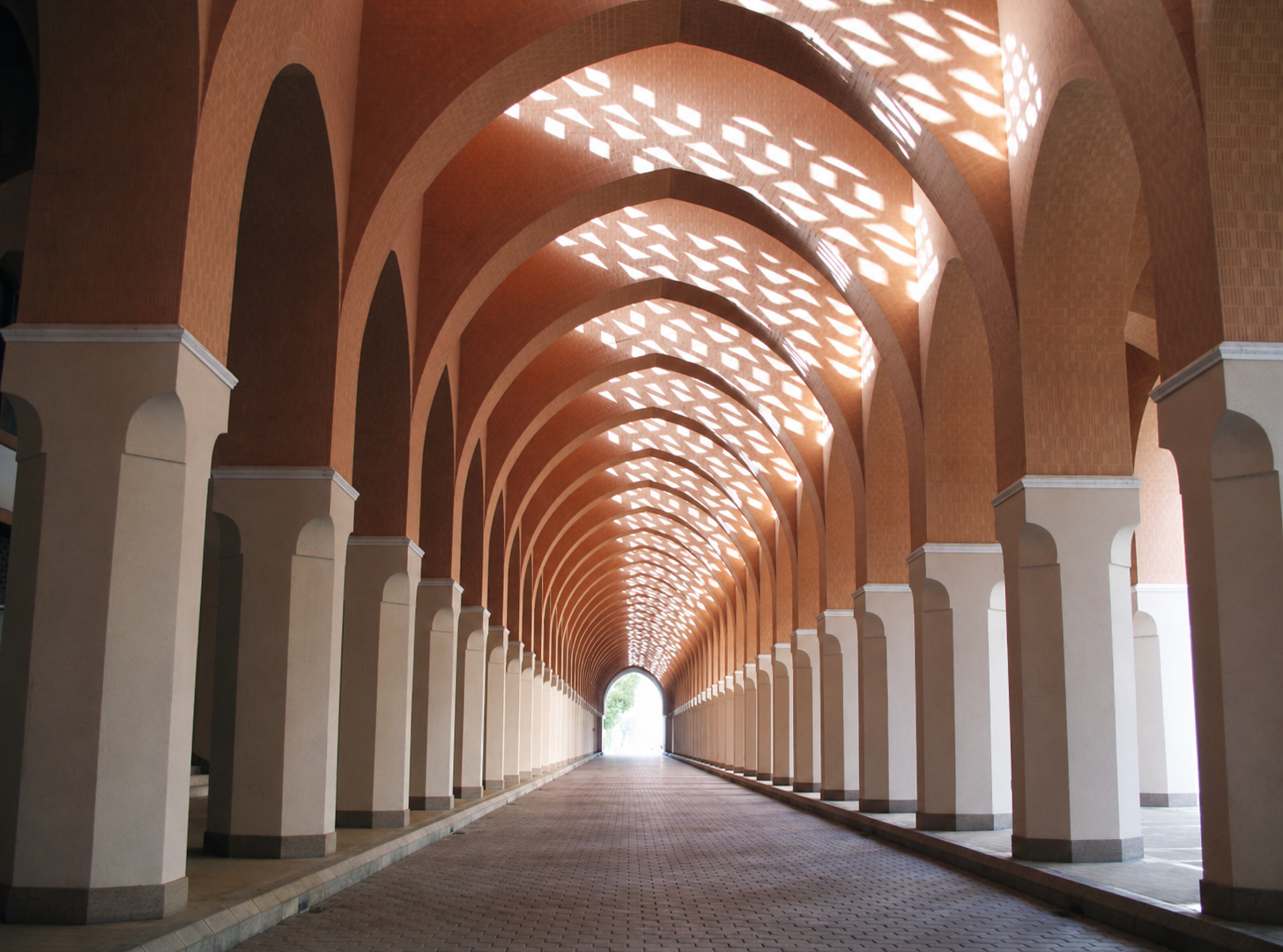 A covered walkway with repeating archways and sunlight creating geometric shadow patterns on the ceiling.