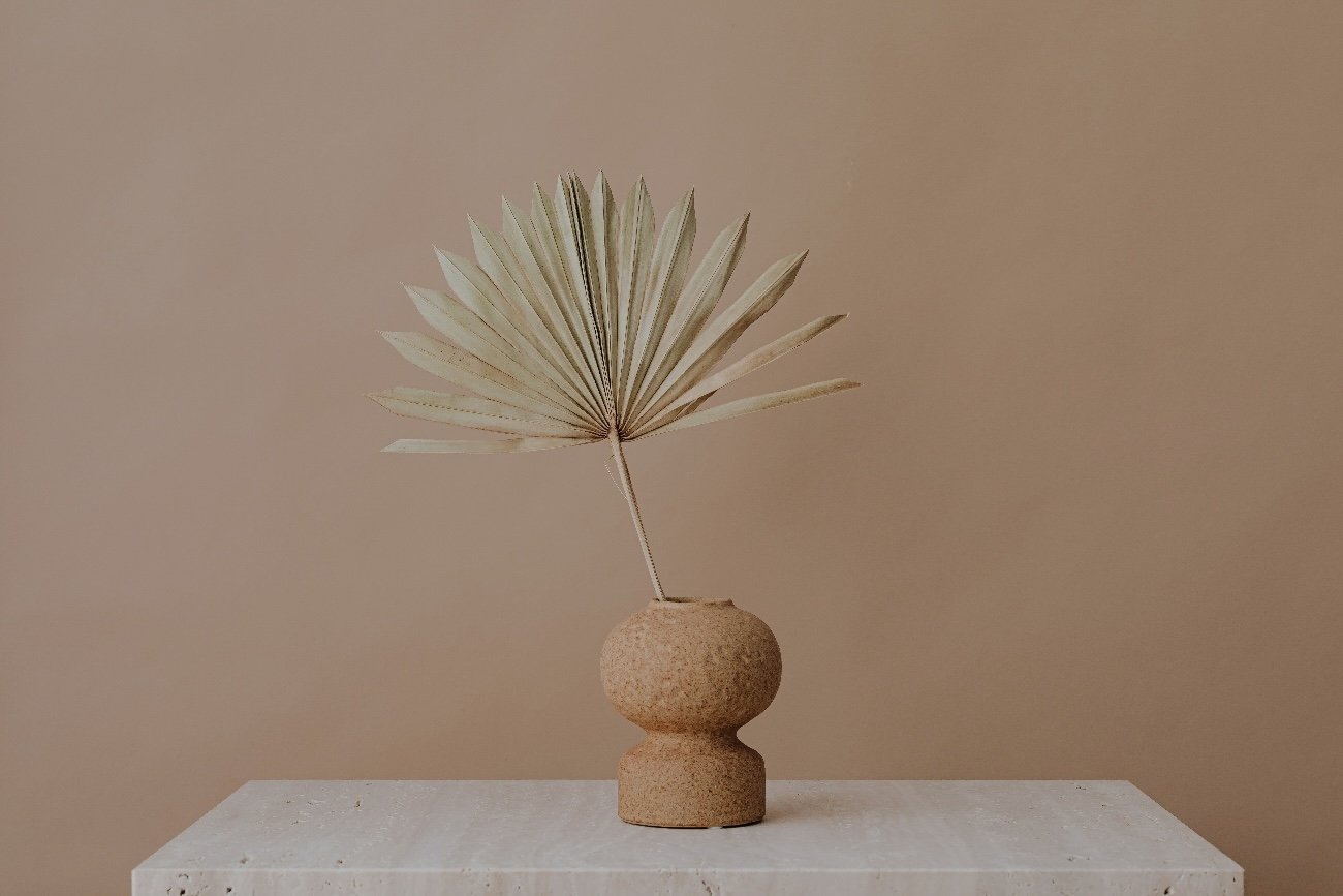 Minimalist beige decorated arrangement with a dried palm leaf in a textured clay vase on a white marble surface.