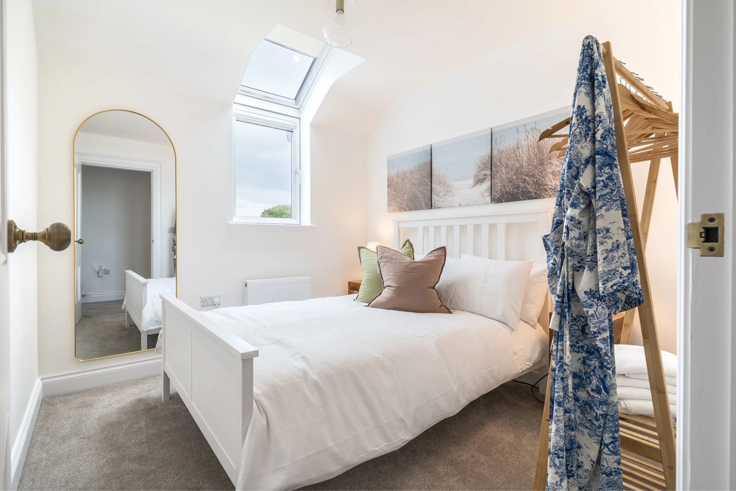 A cozy bedroom with a white bed, green and pink pillows, a large artwork of a beach scene on the wall, a wooden clothing rack with a blue and white patterned dress, a small window with a skylight, and a gold-framed mirror.