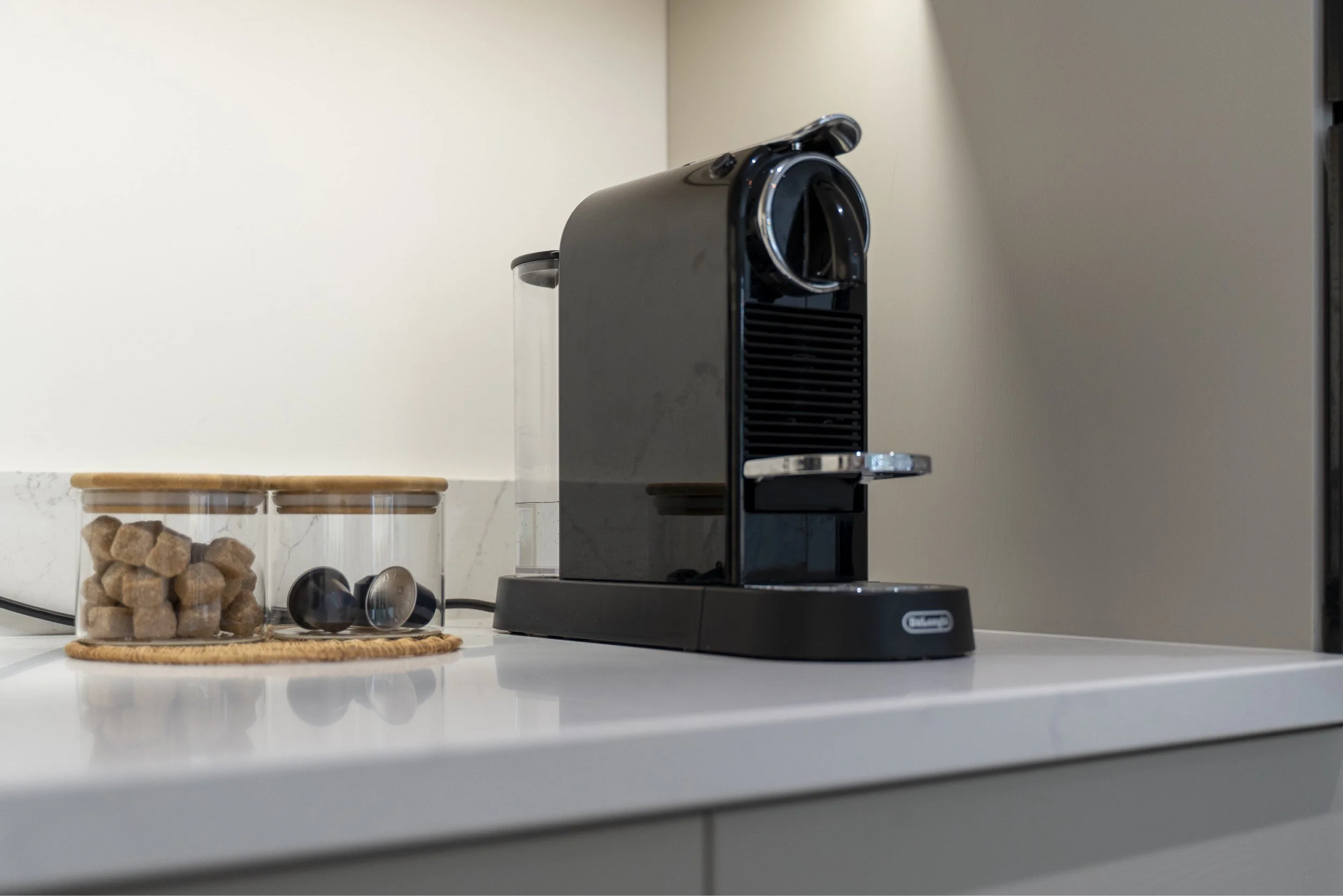 A black Nespresso coffee machine on a white kitchen countertop with a glass jar of brown sugar cubes and a few coffee capsules nearby.
