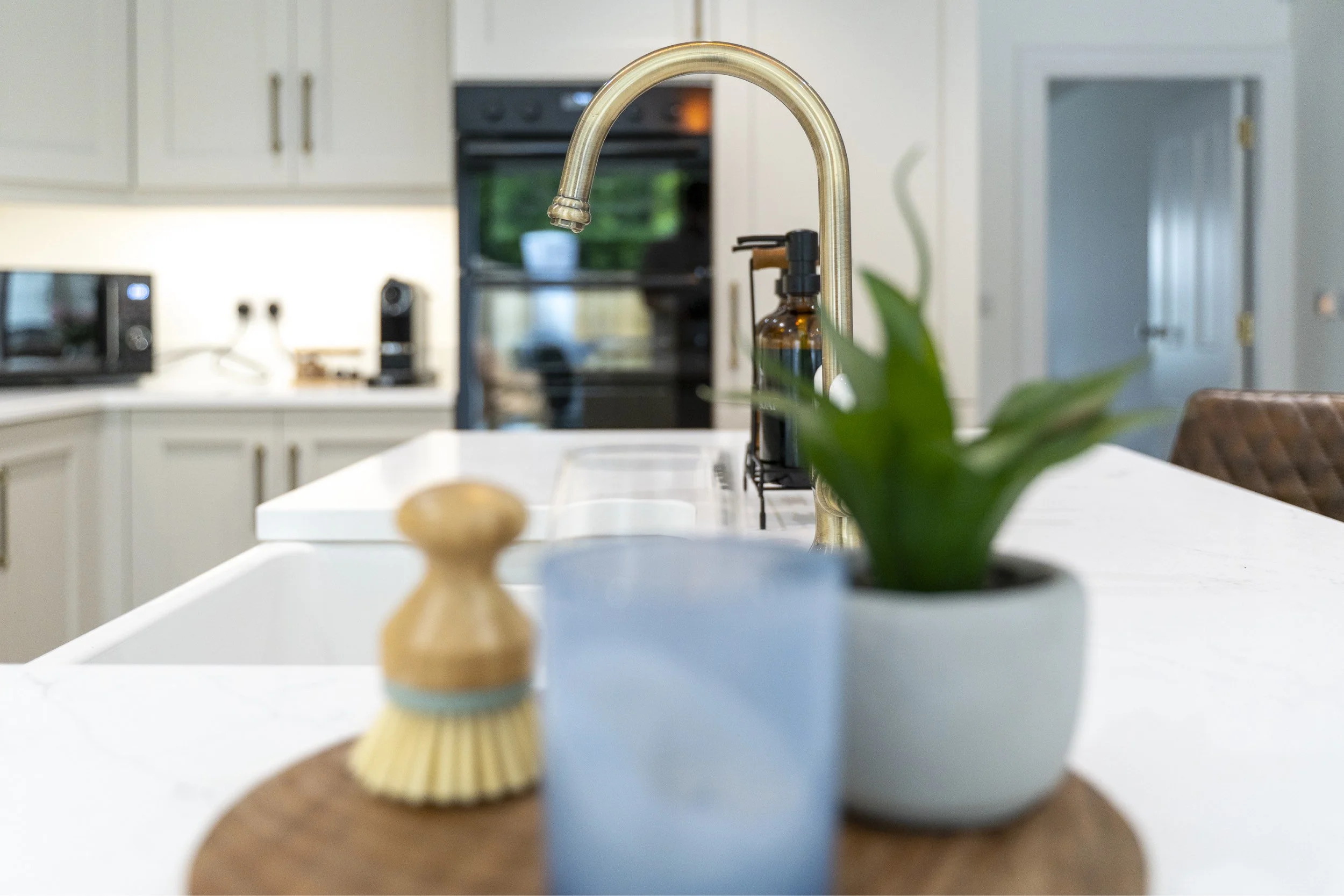 View of a kitchen sink with a gold colored faucet, with a plant and decorative items on the white countertop in the foreground, and kitchen appliances including an oven and microwave in the background.