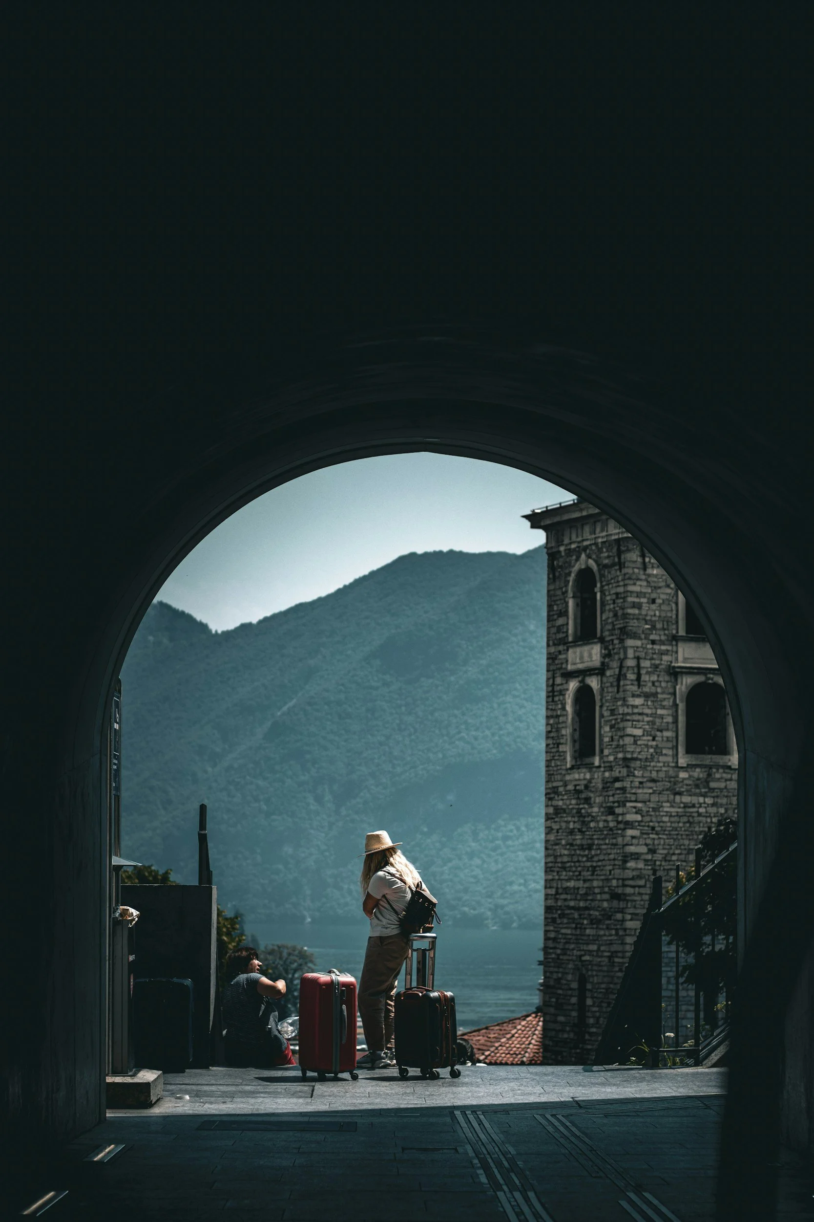 View of travelers waiting with luggage at train station, framed by a tunnel opening, featuring a mountain landscape and a historic building in the background.