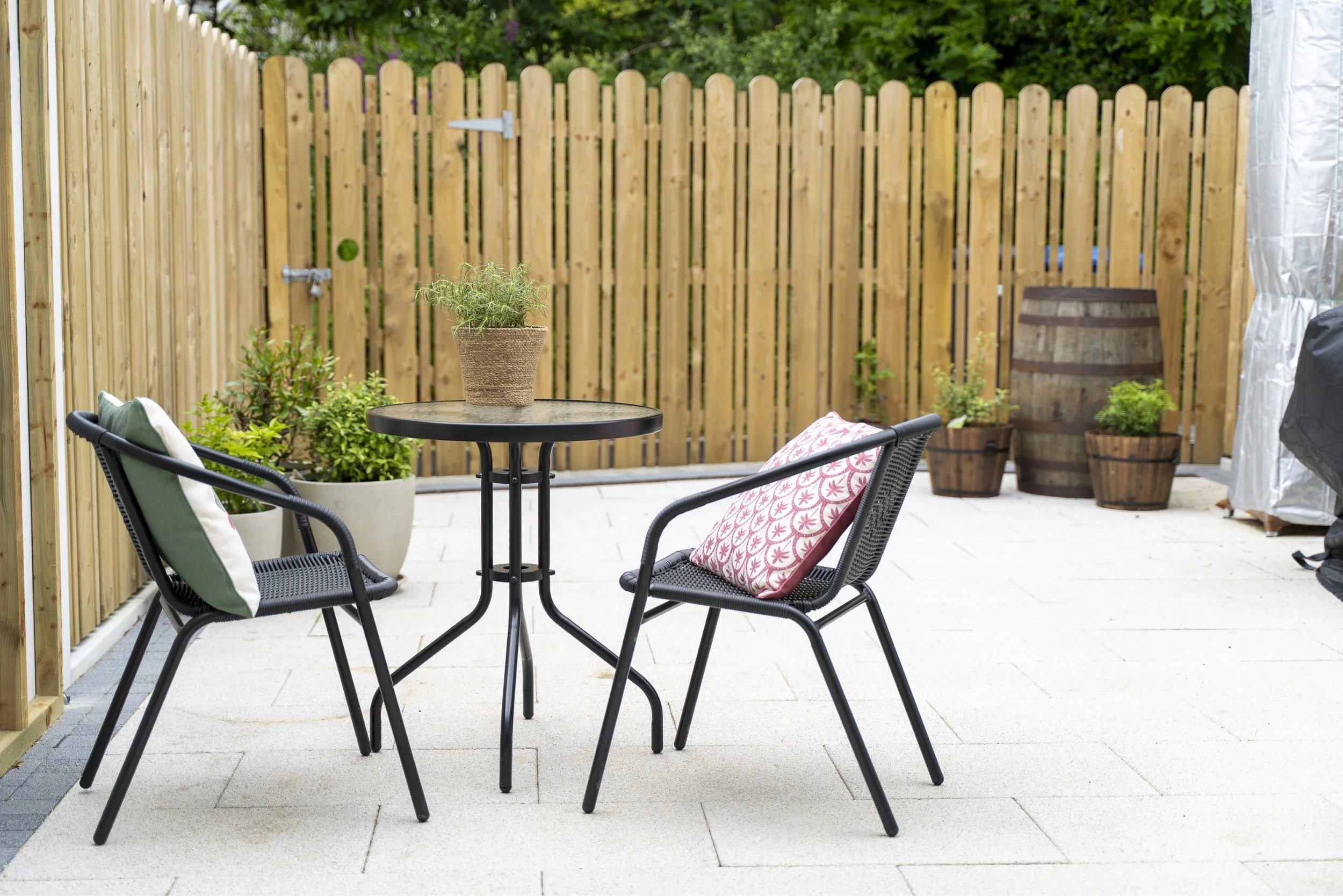 Outdoor patio with a round black table and two black chairs with cushions, surrounded by potted plants and a wooden fence.