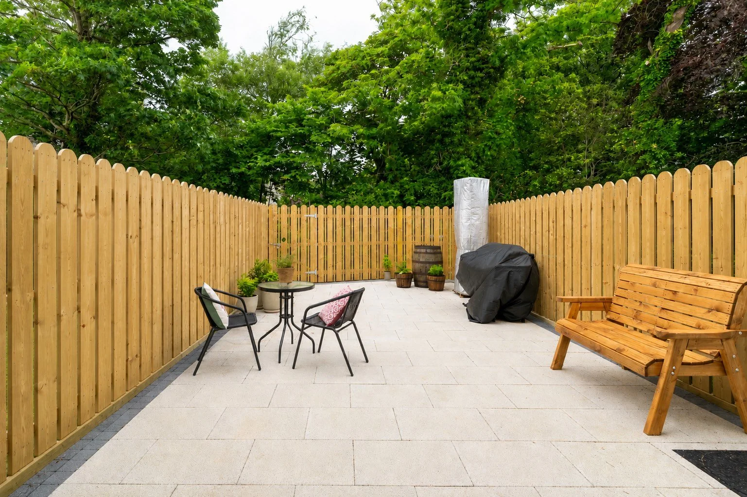Empty backyard patio with wooden fence, black chairs with cushions, small round table, potted plants, a covered grill, a stack of white delivery boxes, and a wooden bench, surrounded by green trees.