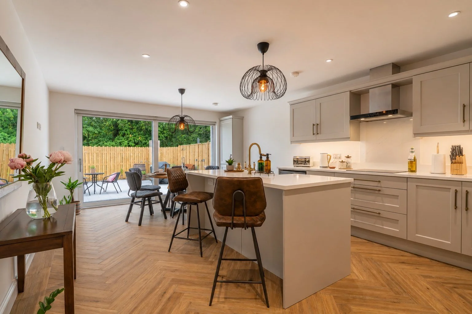 kitchen at The House – Bushmills with modern cabinets, a white island with a sink, wooden bar stools, and a view of a patio with outdoor furniture.