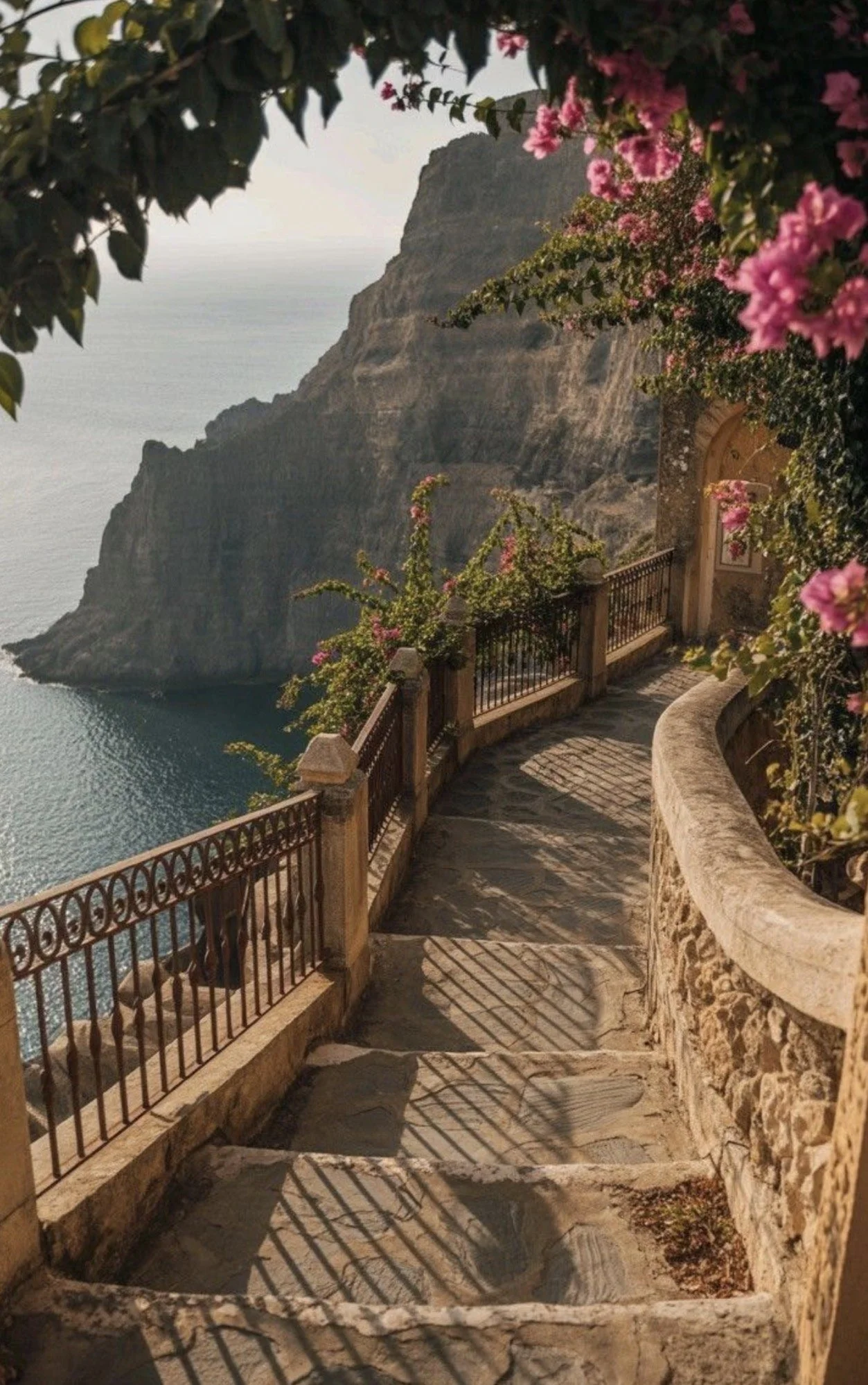 A stone staircase with a metal railing along the edge, leading down beside a stone wall, with flowering bushes and pink blooms overhanging, overlooking a coastal cliff and water.