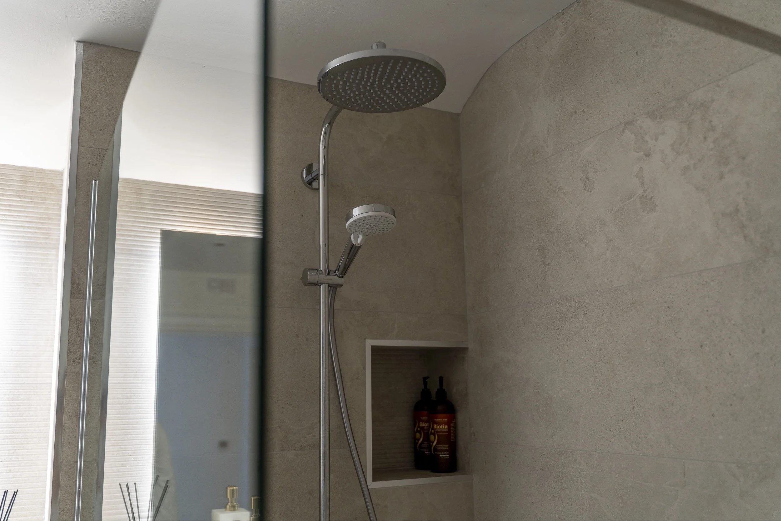Shower area with a round overhead shower head and a smaller handheld shower head, modern beige tiled walls, a built-in shower niche holding two bottles, and a small part of a mirror visible on the left.