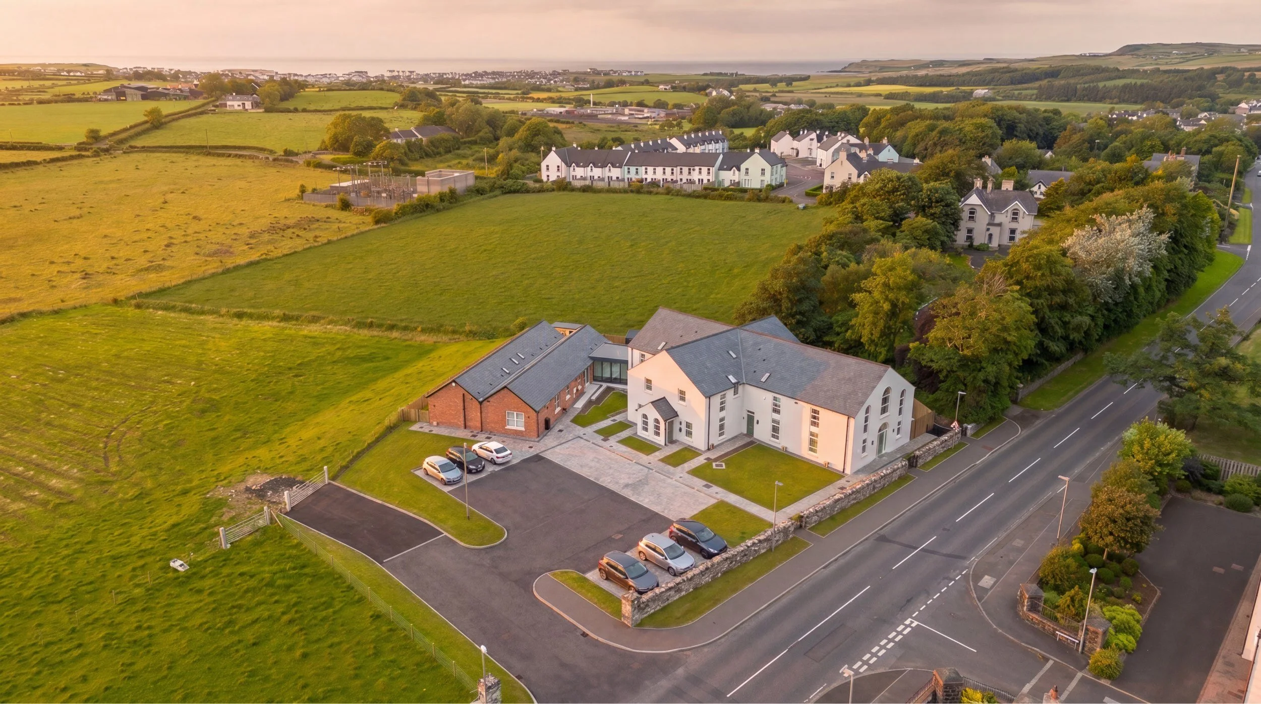 An aerial view of a suburban neighborhood with modern houses, green lawns, and trees, near open fields and the coastline in the background during sunset.