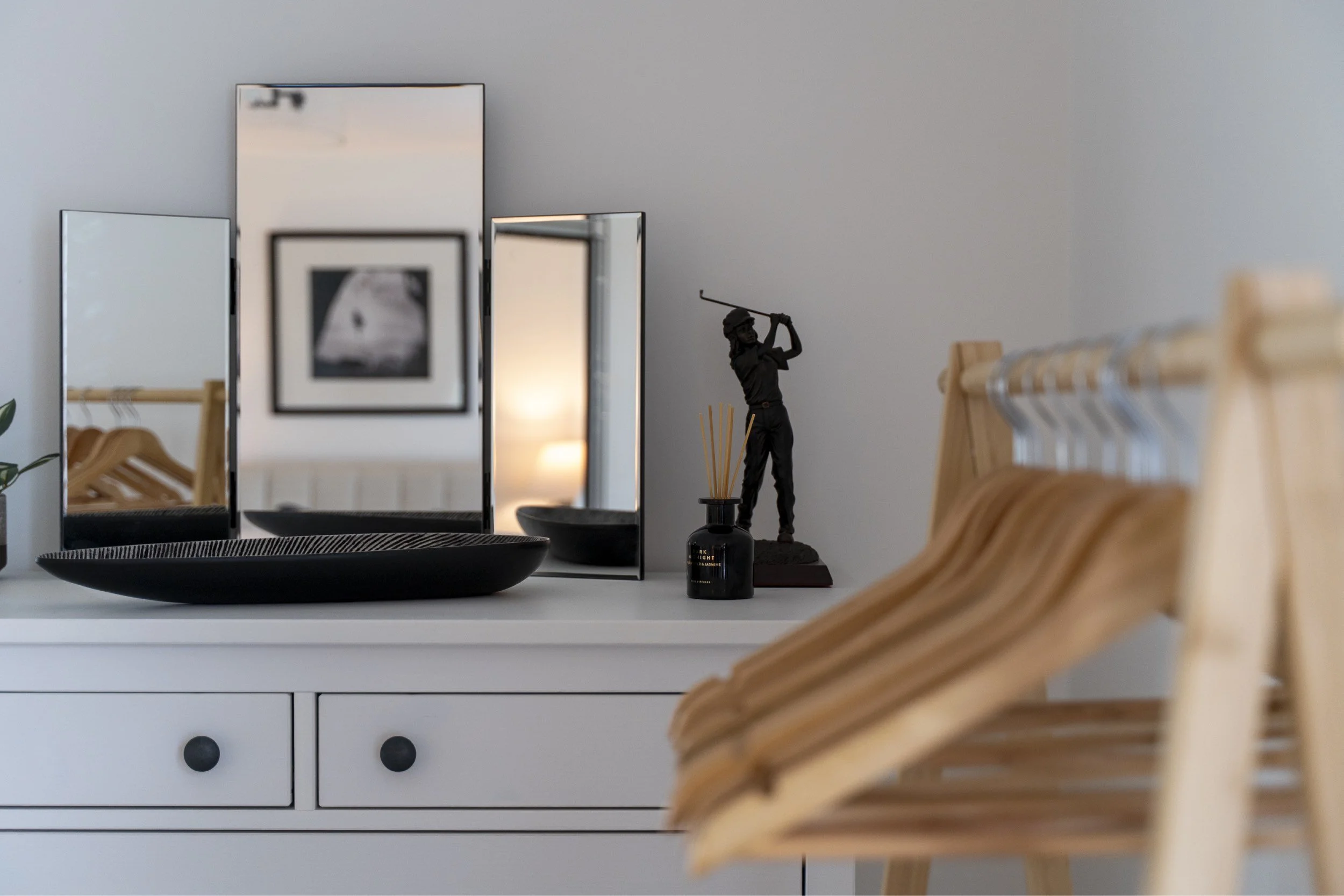 A white dresser with a black tray on top, three mirrors, a black candle holder with a woman figurine holding a golf club, and wooden coat hangers hanging on a rack to the right.