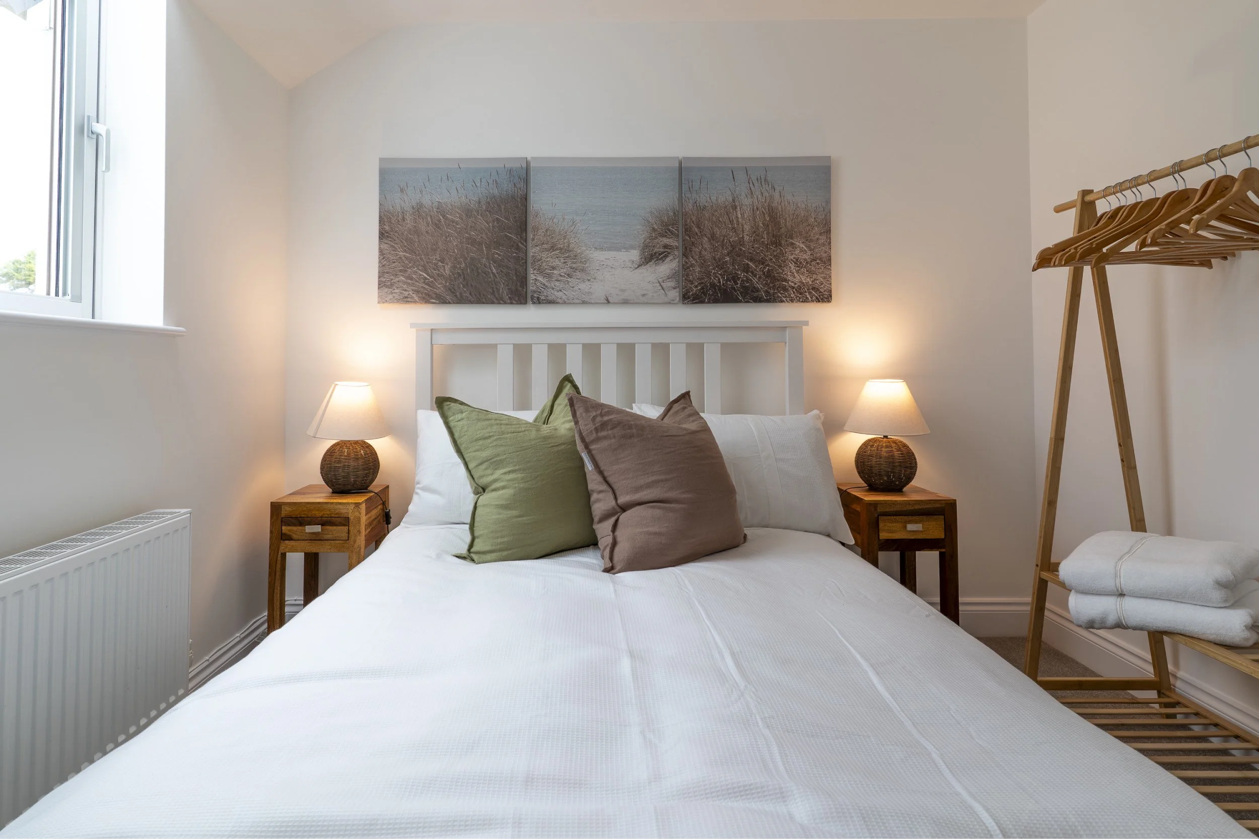 A bedroom with a white bed, green and brown pillows, wooden nightstands with lamps, a triptych beach art piece above the bed, a window on the left, and a wooden clothes rack on the right.