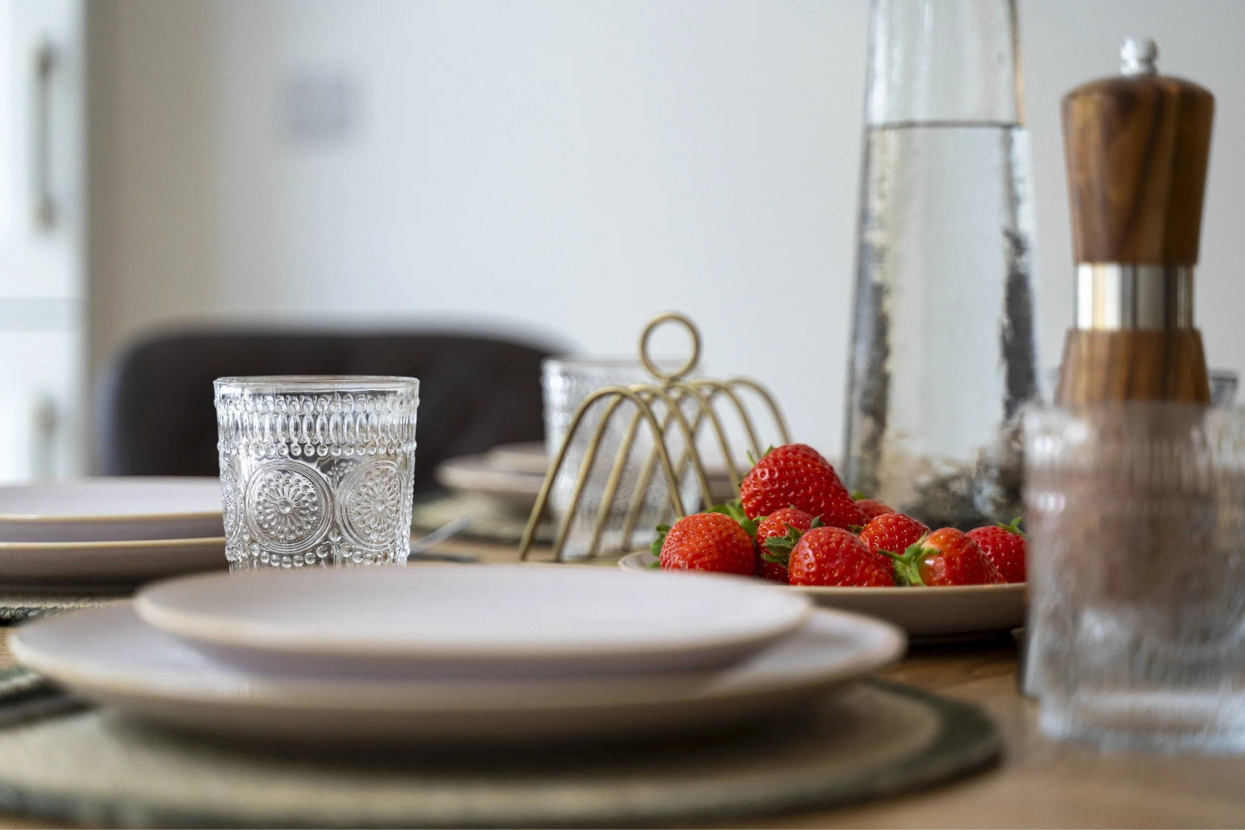 A dining table set with glassware, white plates, and a bowl of fresh strawberries, with blurred background elements including a water carafe and a pepper grinder.