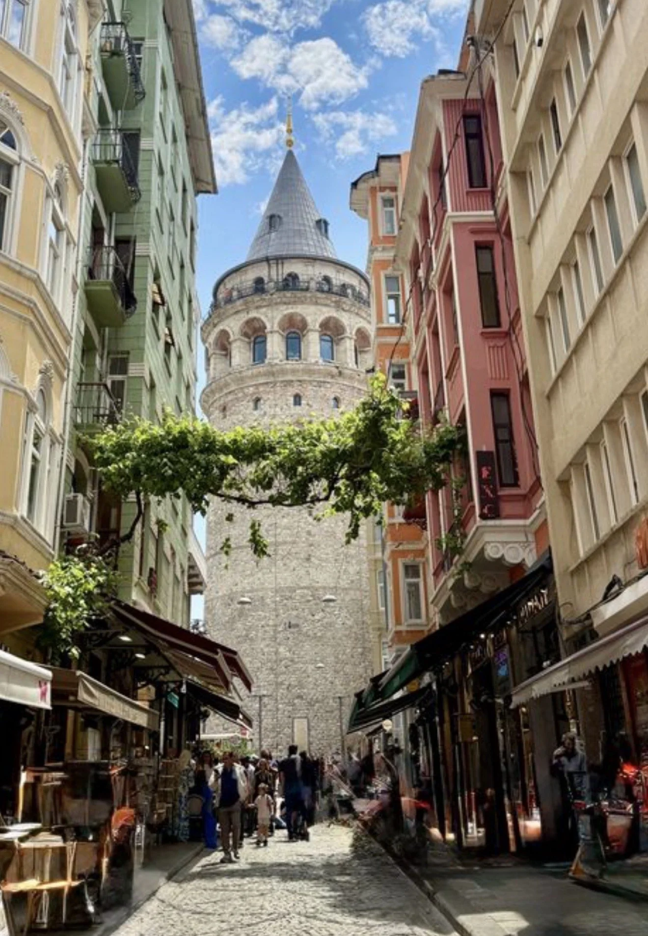 A bustling street scene in Istanbul, Turkey, with the historic Galata Tower in the background, flanked by colorful buildings, shops, and cafes, under a blue sky with clouds.