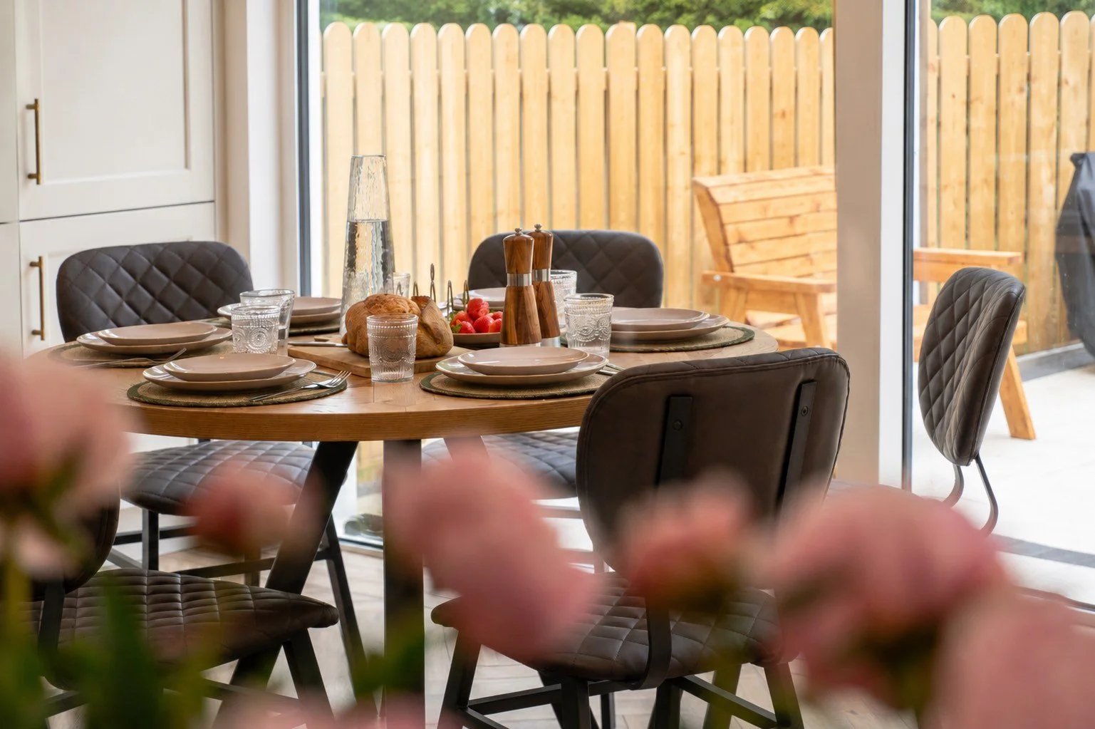 A dining table set for five people with plates, glasses, and cutlery, decorated with a bread and a bowl of strawberries in a sunlit room with a wooden fence visible outside.