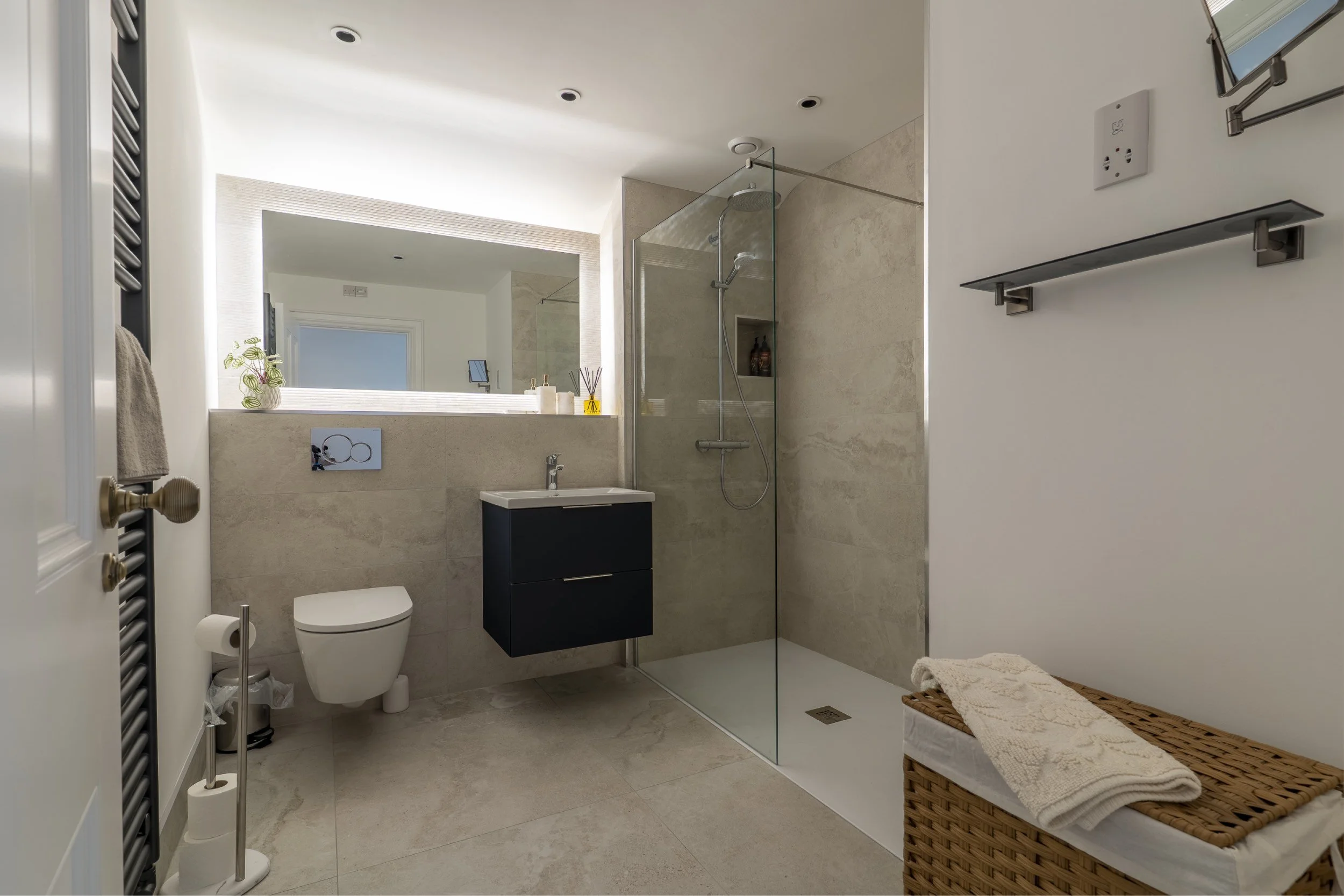 Modern bathroom with beige tiled walls and floor, glass shower enclosure, wall-mounted toilet, vanity with dark cabinet, large mirror, potted plant, and wicker basket with towels.