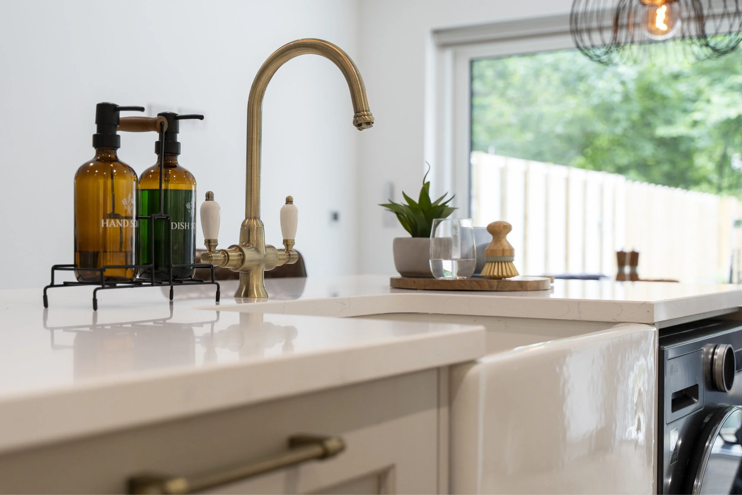 A modern kitchen countertop with a gold faucet, soap dispensers, a small potted plant, a glass of water, a wooden scrub brush, and a soap dish, with a large window showing trees outside.