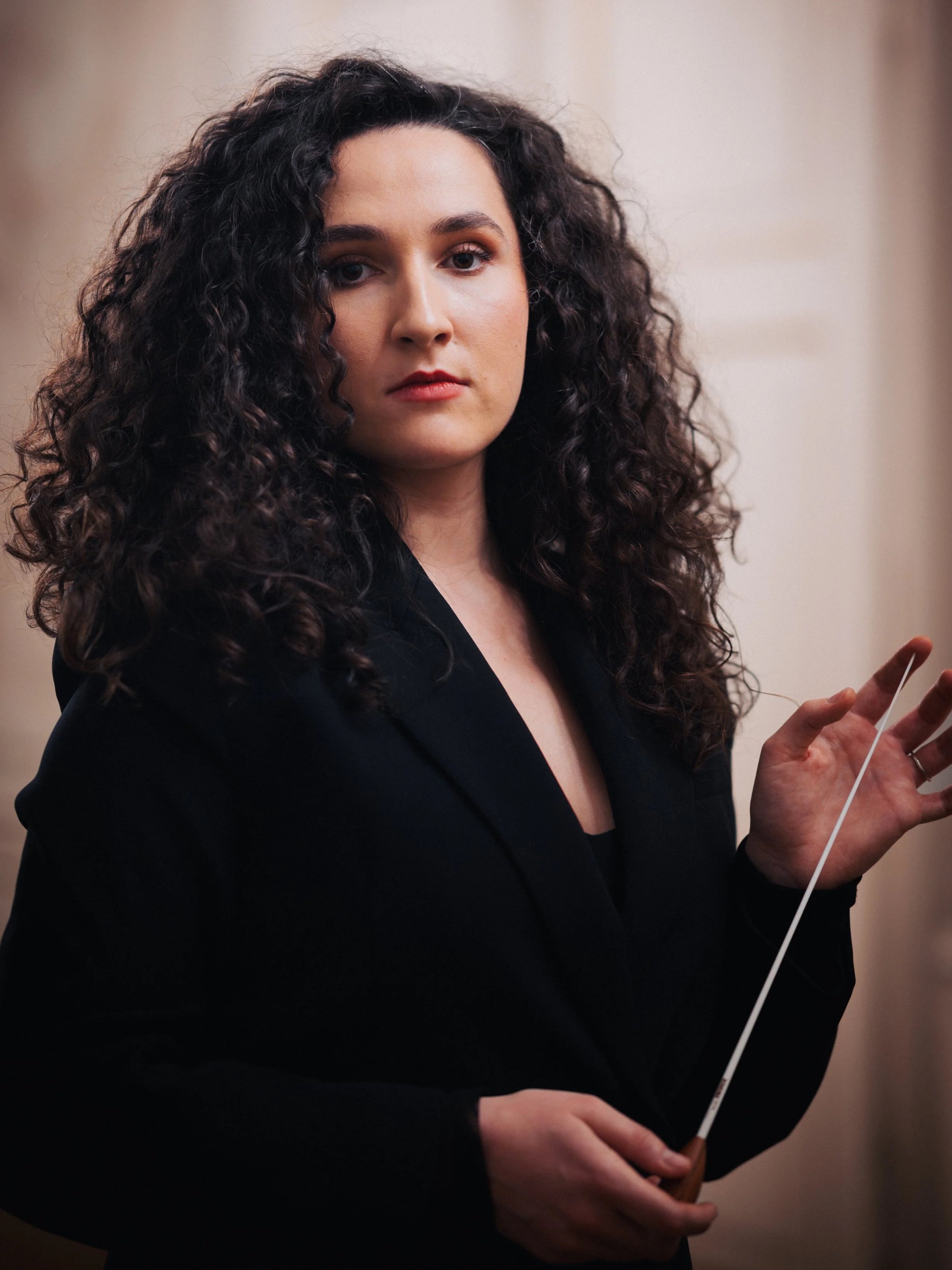 A woman with curly dark hair dressed in black, holding a white object, stands against a neutral background.
