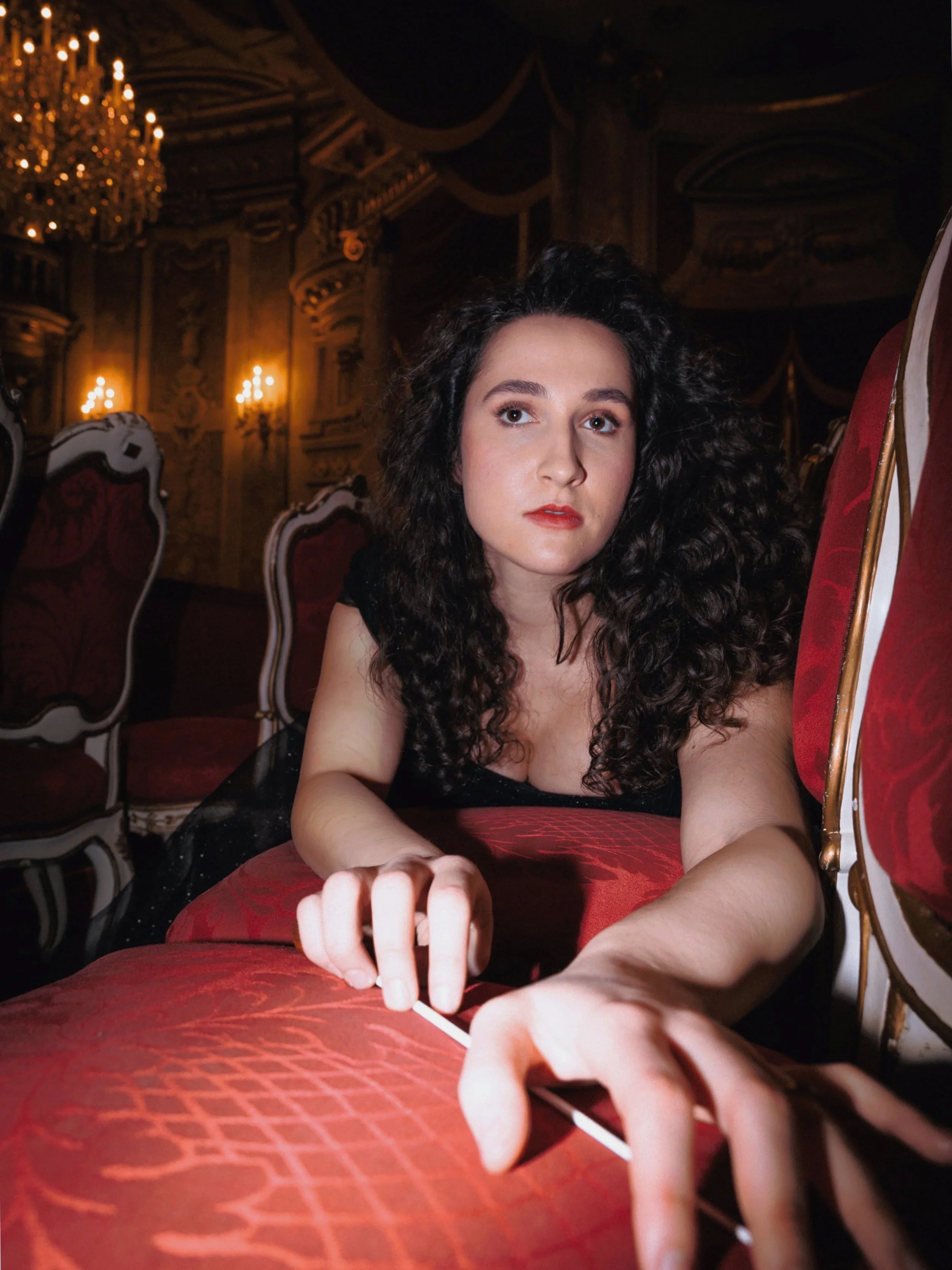 A singer-conductor with curly dark hair and red lipstick sitting in an ornate, dimly lit theatre with red velvet chairs and a chandelier, reaching her baton.