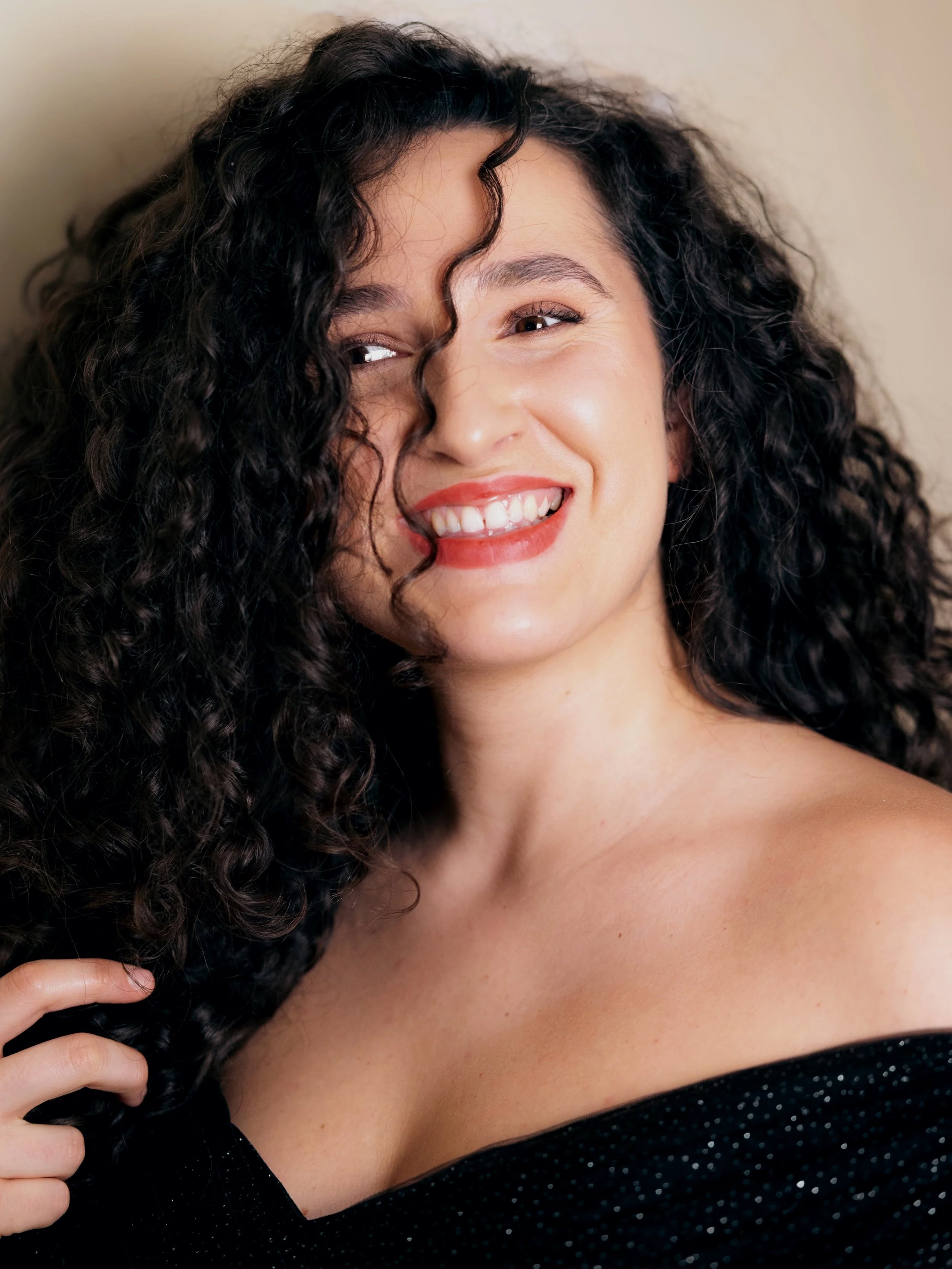 Close-up of a smiling woman with long curly black hair, wearing a black off-shoulder top with small white polka dots, against a beige background.