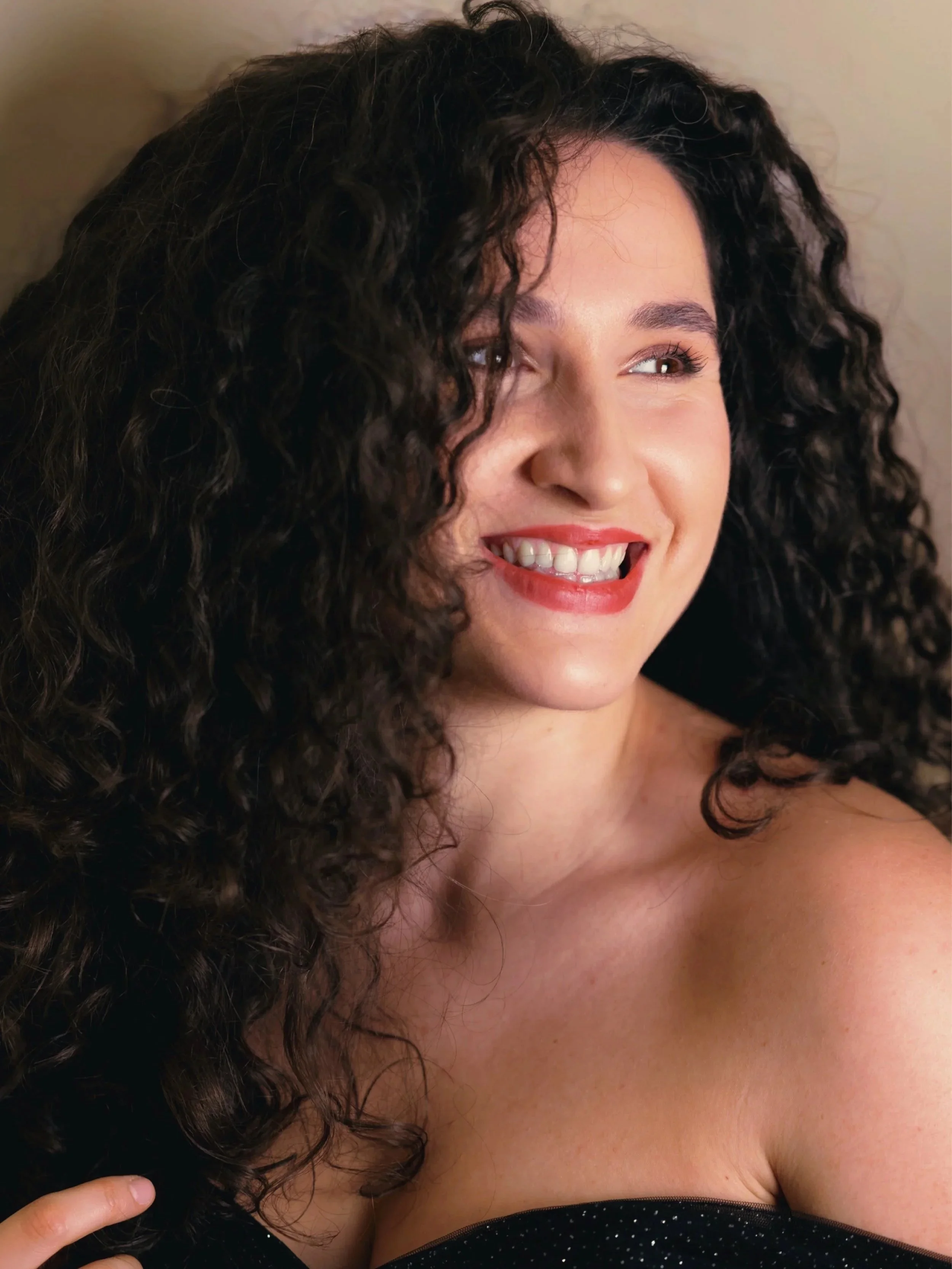 Close-up of a woman with curly dark hair smiling, showing teeth, wearing a black top with small white dots.