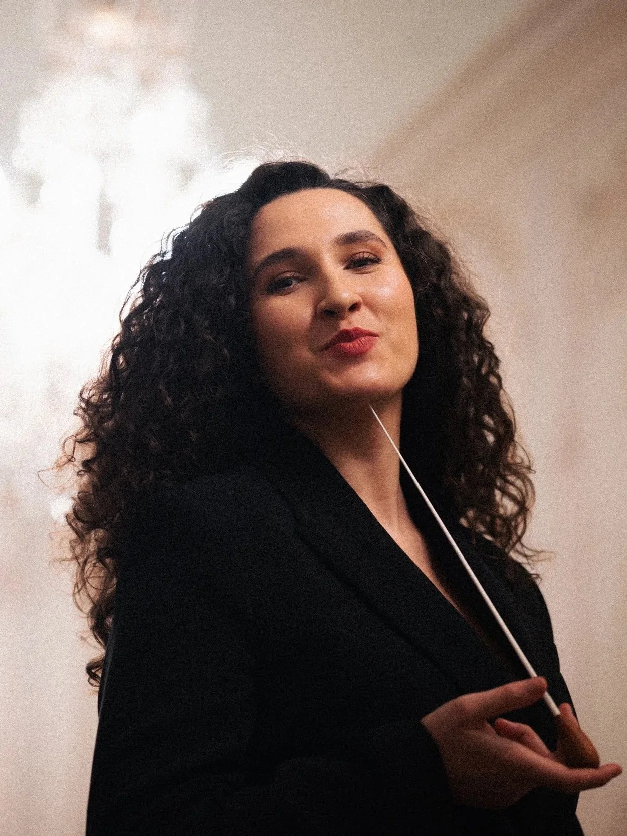 A conductor with curly dark hair, wearing a black blazer, holding a white baton in her hand, posing confidently against a neutral background.