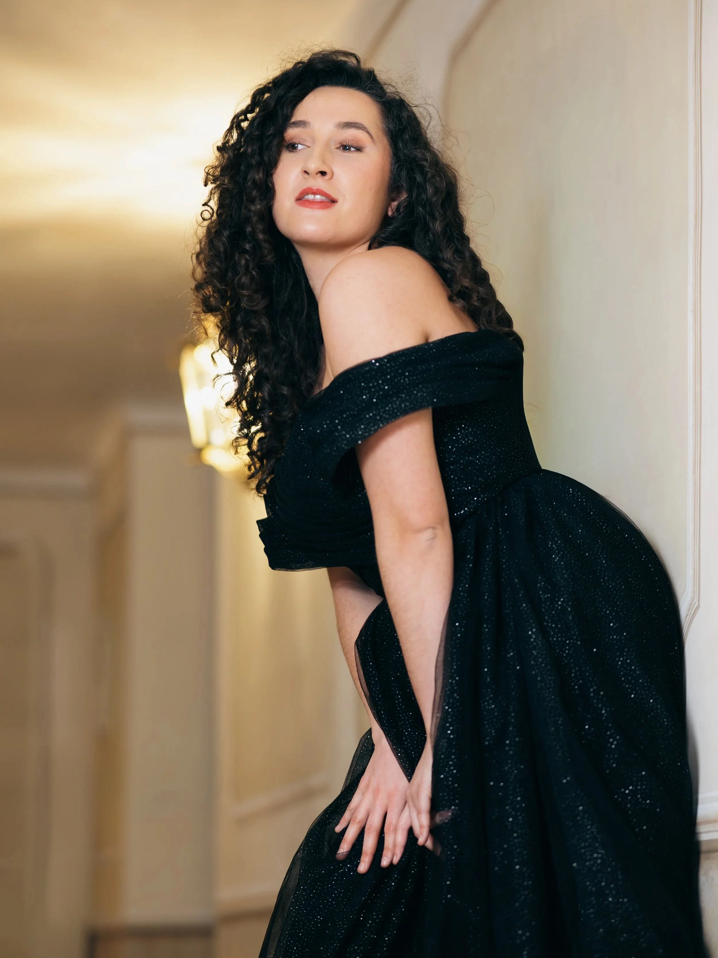 A woman with long curly dark hair wearing an off-the-shoulder black dress with glitter details, standing against a beige wall with soft lighting.